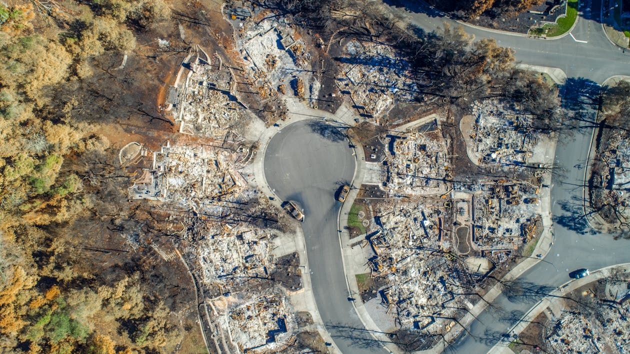 Aerial view of a neighborhood destroyed by fire, showing burned houses, a road with cars, and surrounding charred trees.