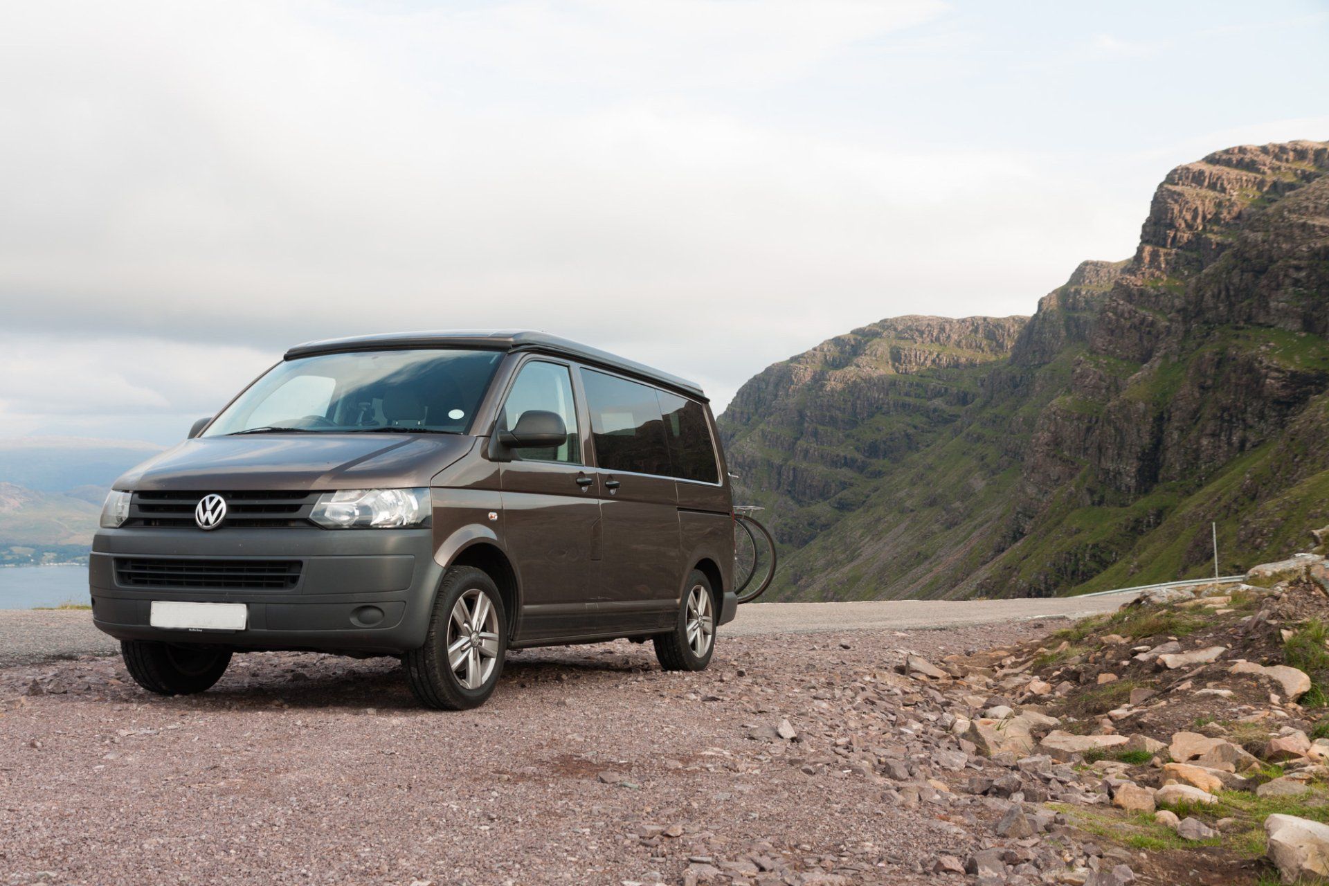 Brown VW campervan parked on a gravel road with mountains in the background.