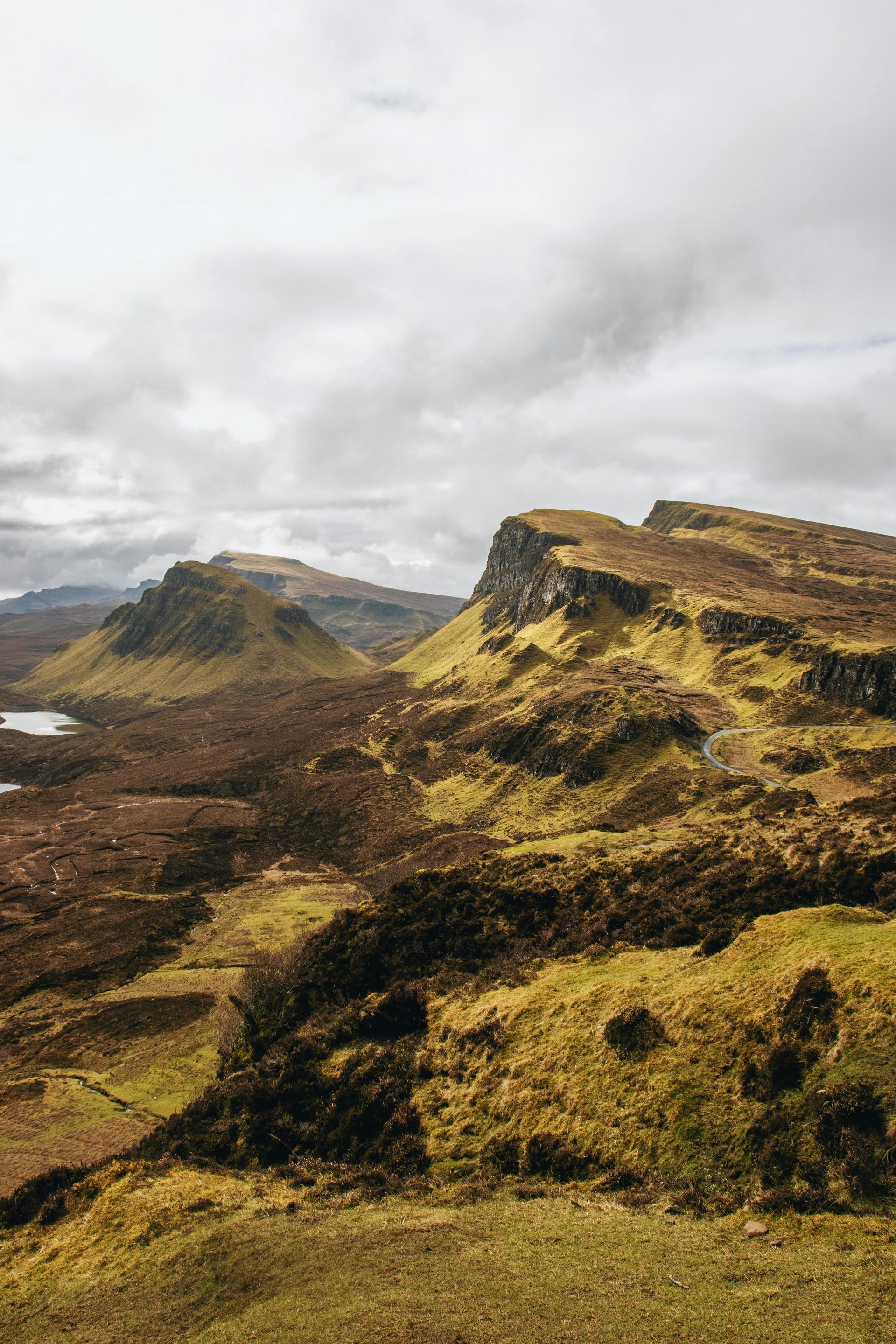 Rolling Scottish highlands under a cloudy sky, featuring rugged terrain and a small lake.
