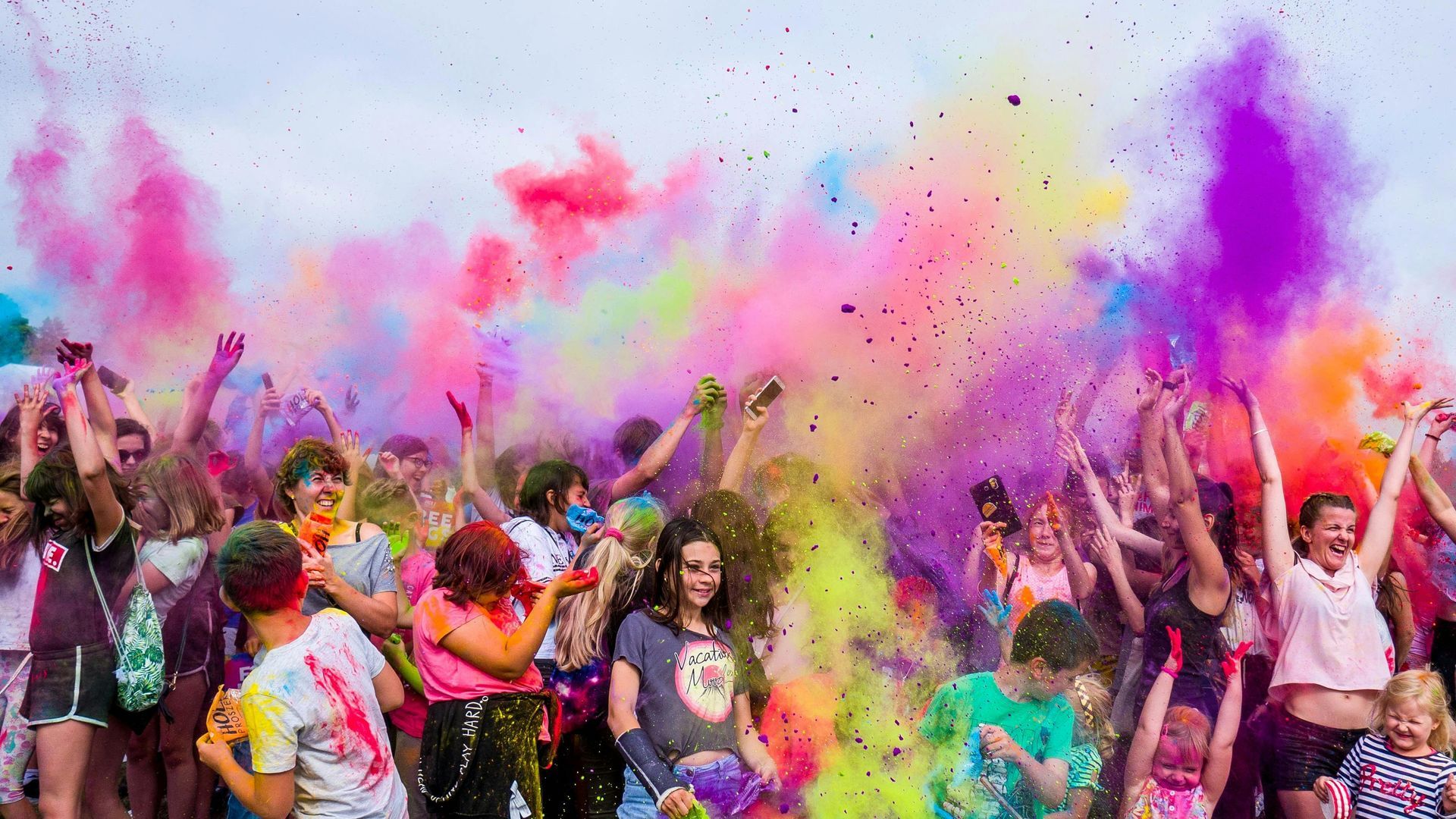 People celebrating outdoors, throwing colorful powder in the air. Bright pink, purple, yellow, green hues.