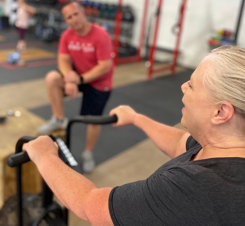 A woman is riding an exercise bike in a gym while a man sits in the background.