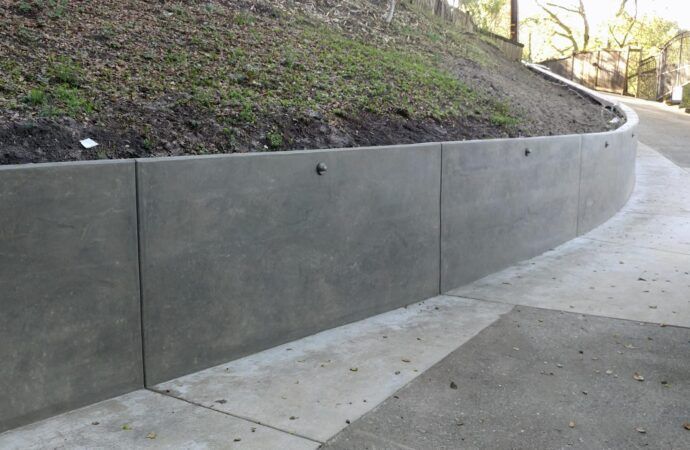 Concrete retaining wall curving alongside a paved walkway, with a grassy hillside above.
