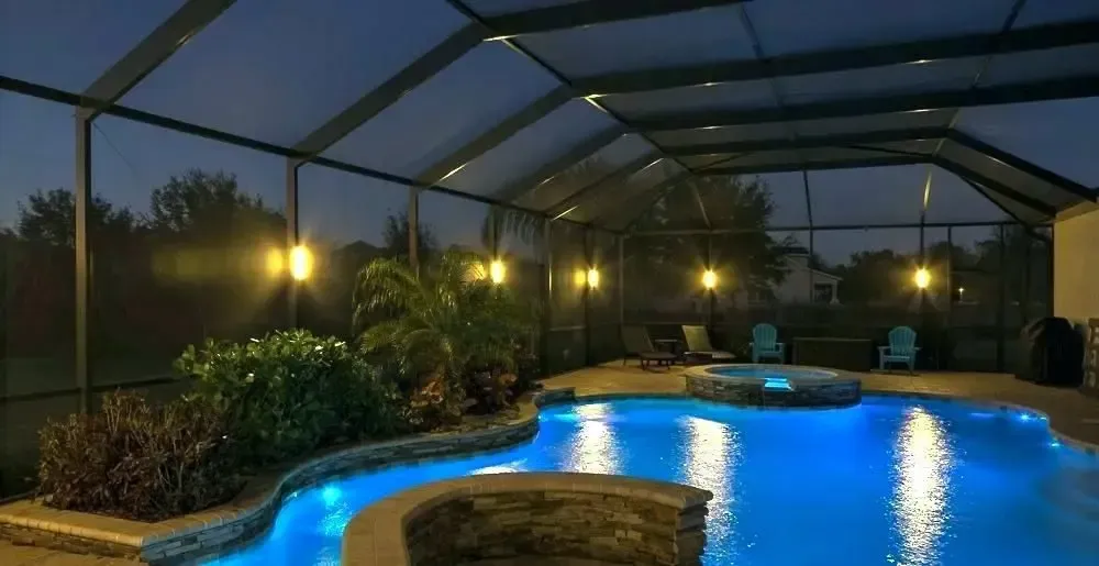 Nighttime pool scene with illuminated blue water, screened enclosure, and potted plants.