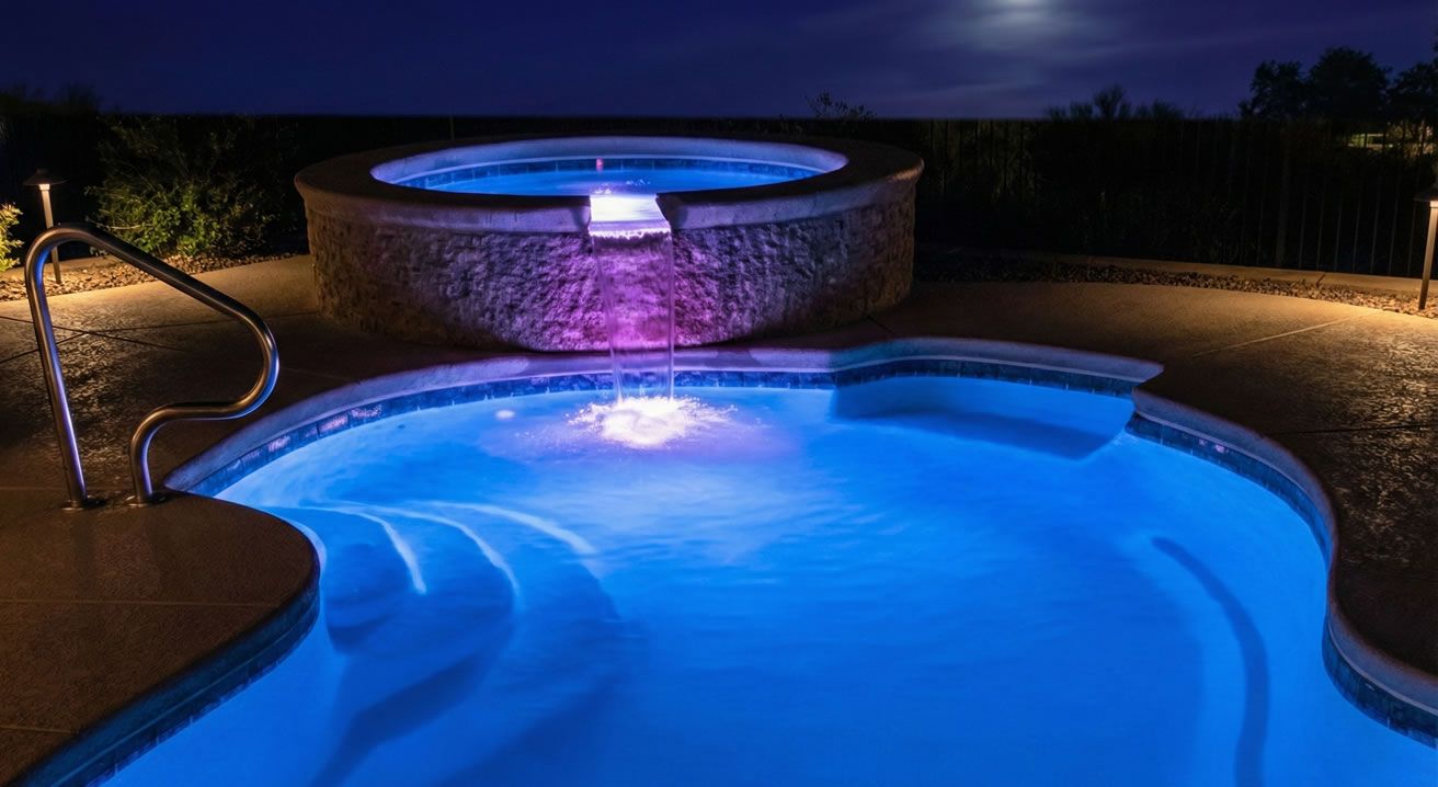Pool and hot tub illuminated at night, with blue and purple underwater lights.