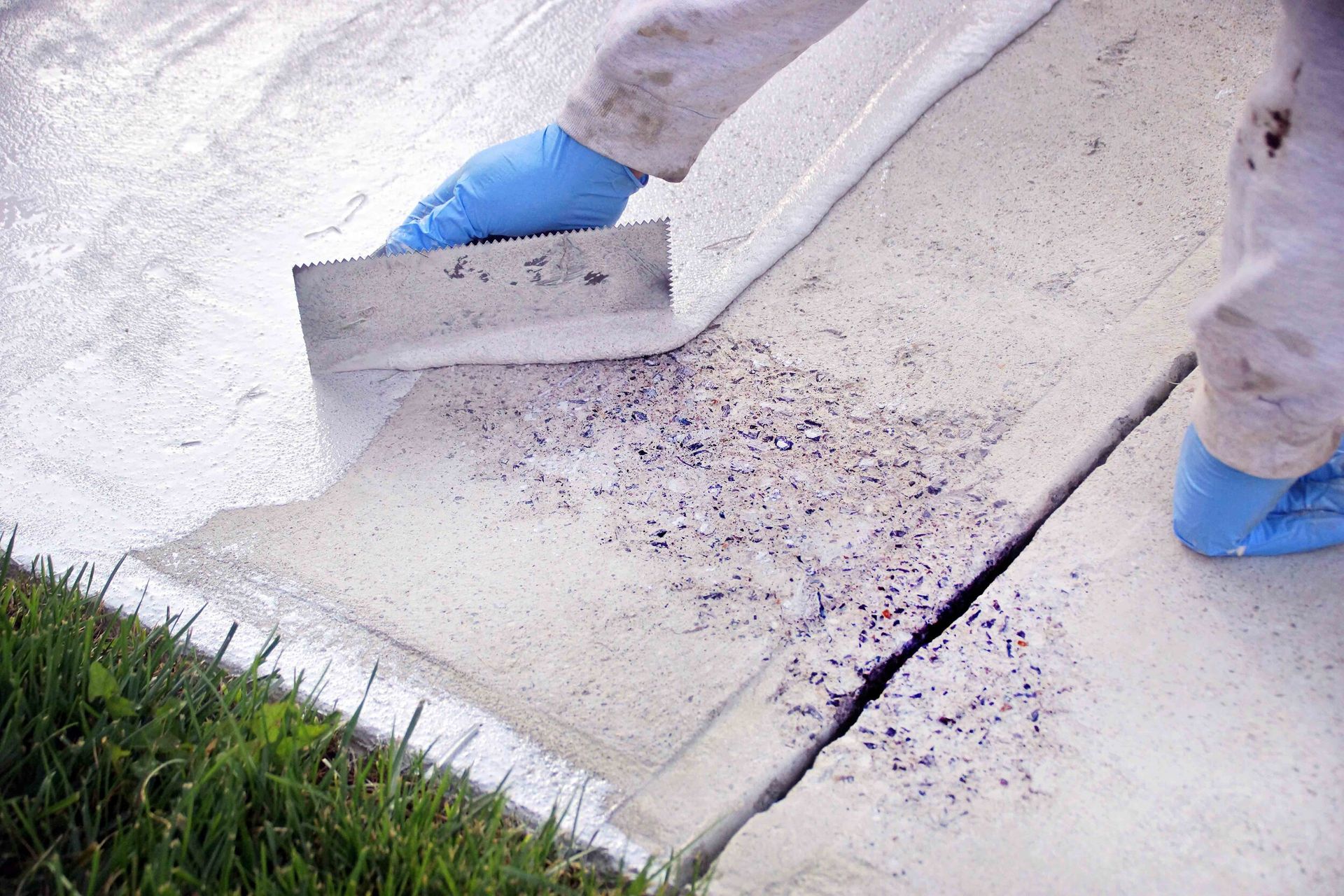 Person applying white sealant to a cracked concrete surface using a trowel, wearing blue gloves.