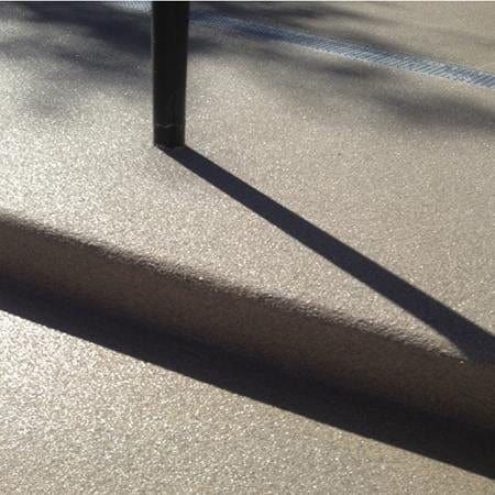 Shadow of a pole on textured, light-colored ground. Sunlight casts a dark rectangle.