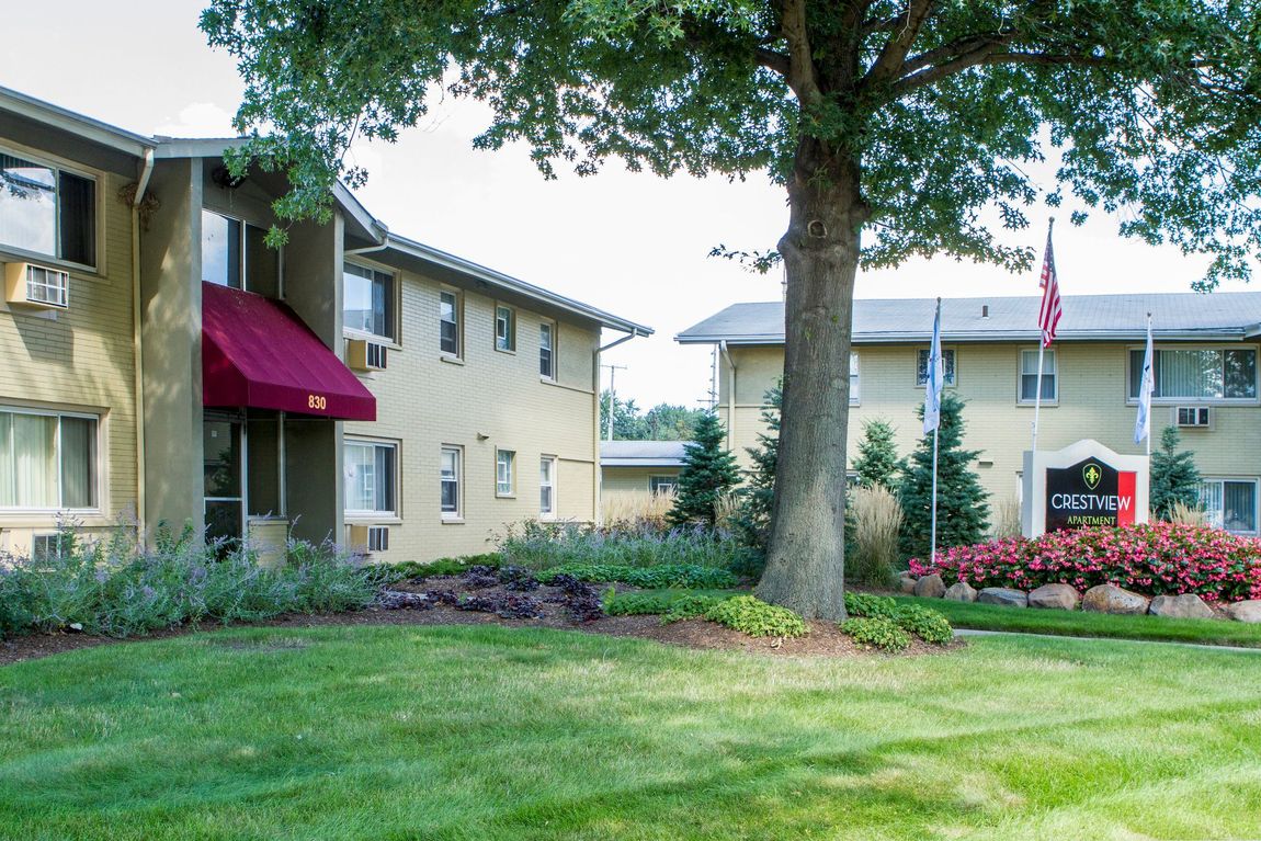 Apartment buildings with green lawn, shrubs, and trees. Red awning over door. Flags fly in the distance.