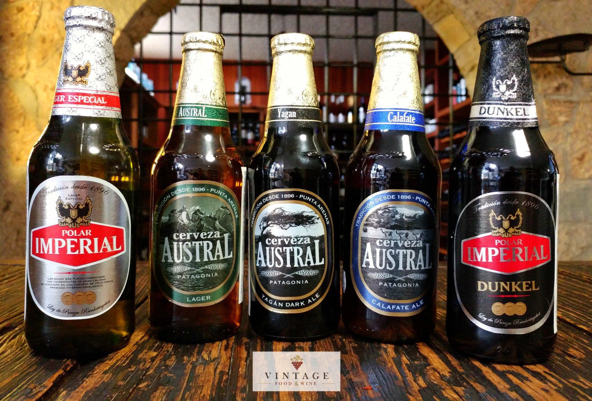 Cinco botellas de cerveza Imperial Austral están alineadas sobre una mesa de madera.