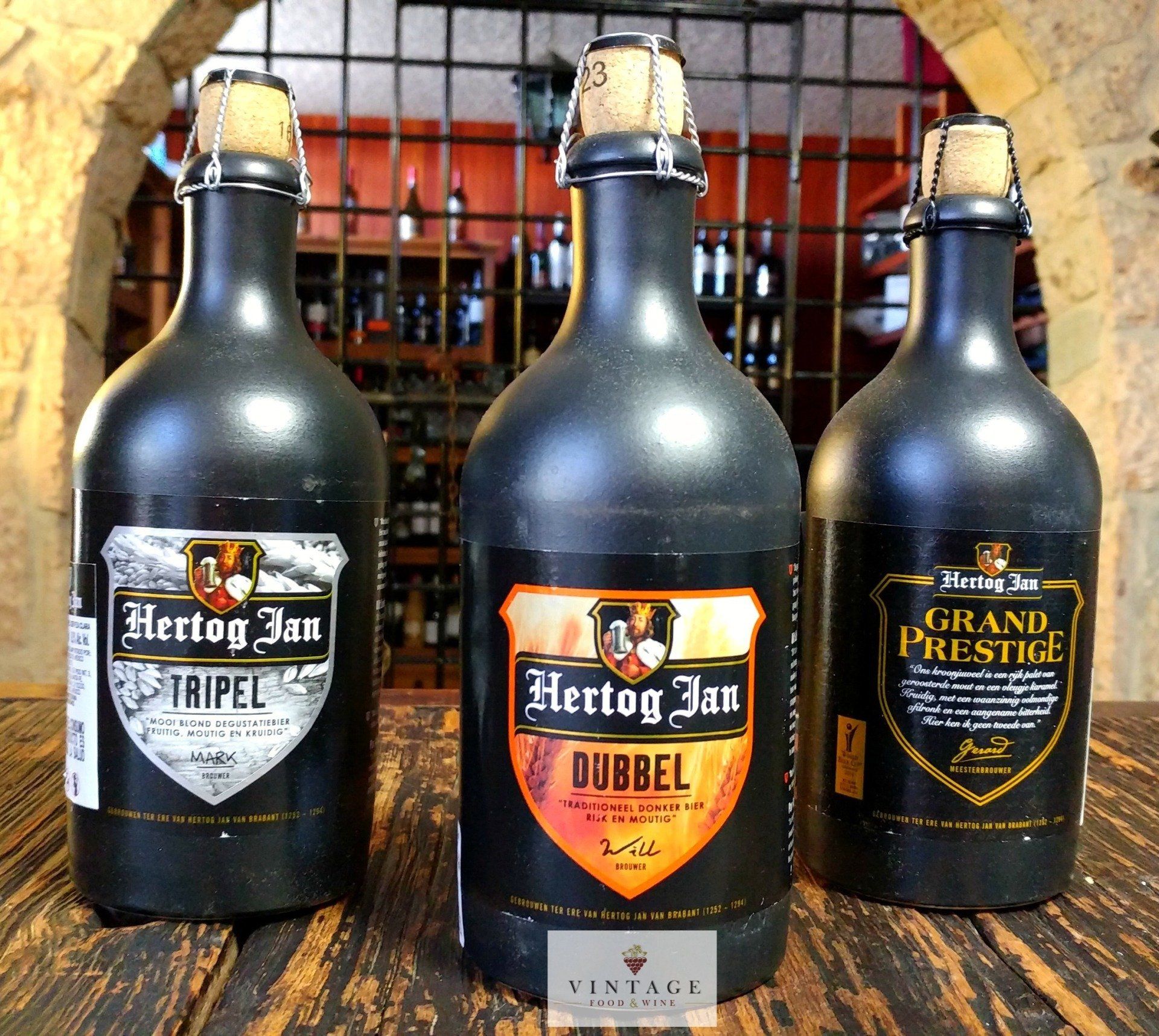 Tres botellas de Hertog Jan están alineadas sobre una mesa de madera.