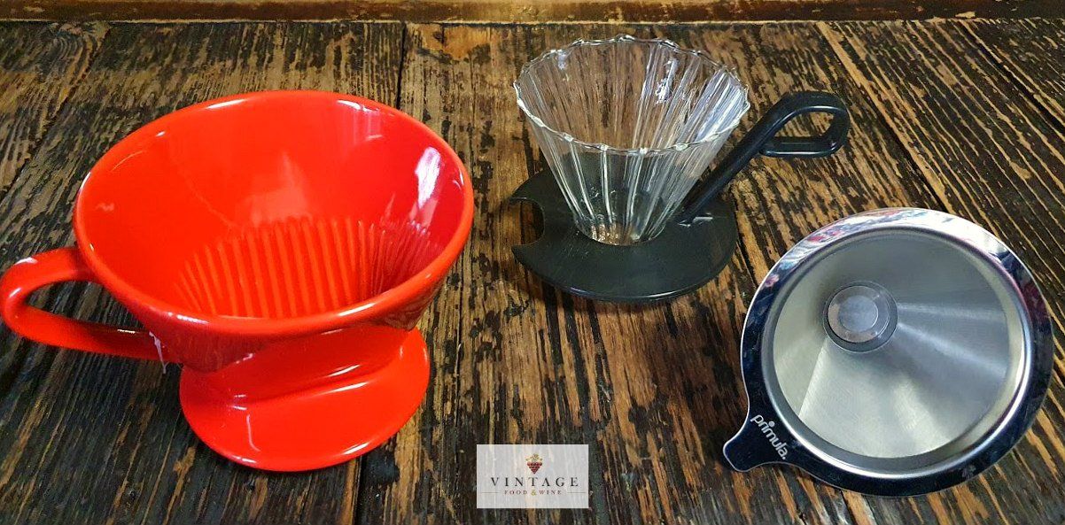 Una taza roja, una taza de cristal y un embudo de metal están sobre una mesa de madera.