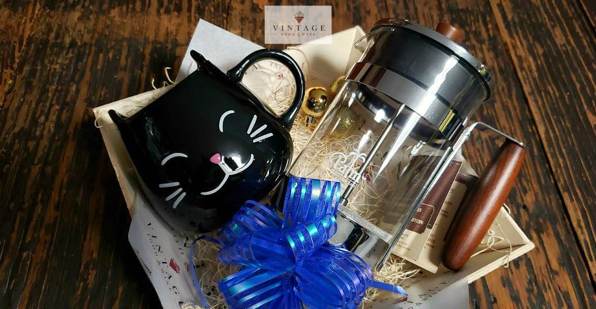 Una canasta de regalo con una taza de gato y una prensa francesa sobre una mesa de madera.