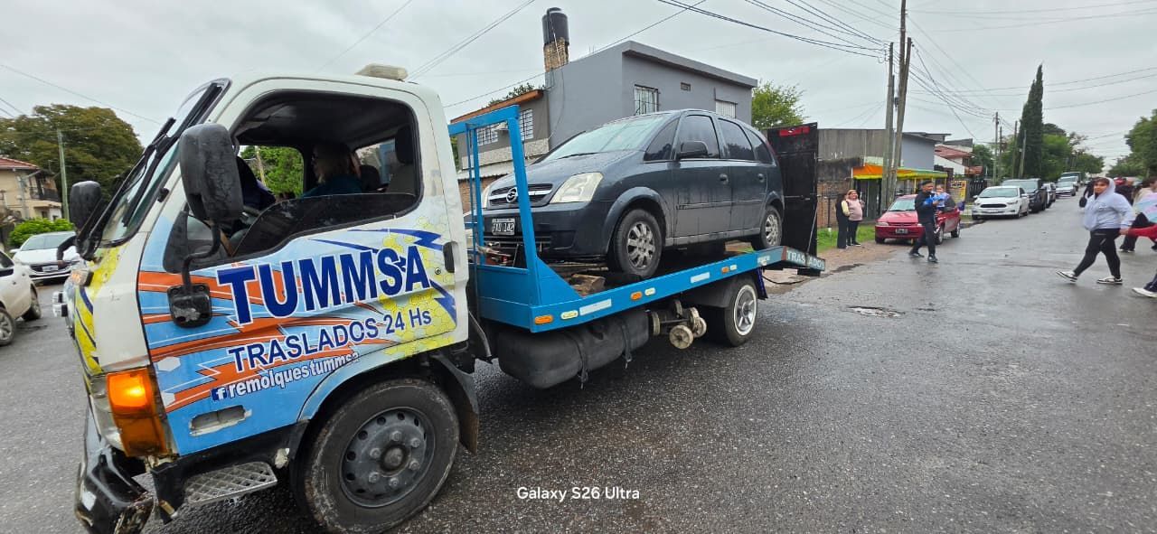 Una grúa remolca un SUV gris en una calle mojada de la ciudad, con gente cerca y un cielo nublado.