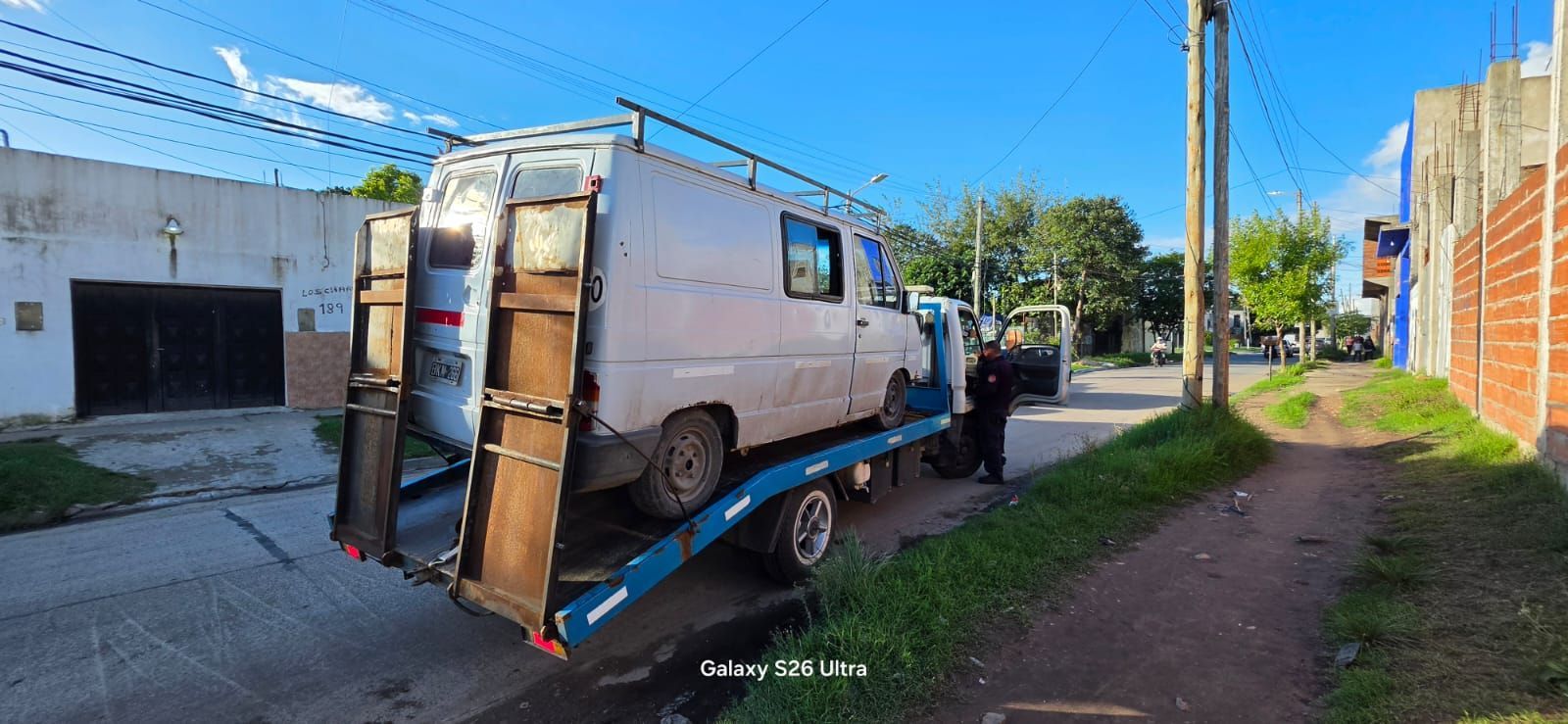 Un camión volquete con rampa trasera estacionado en una calle soleada junto a casas y una persona de pie cerca.