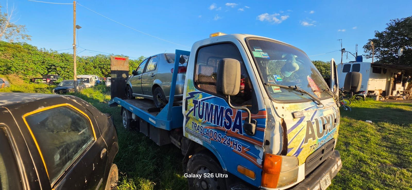 Una grúa transporta un coche negro, con un camión de servicio azul estacionado en una carretera soleada.