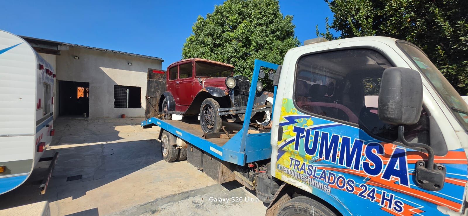 Una grúa transporta un coche rojo en una entrada soleada junto a un garaje y un árbol.