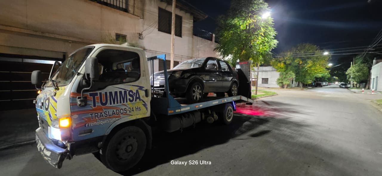 Grúa de remolque nocturna transportando un coche dañado en una calle de la ciudad cerca de edificios y árboles.