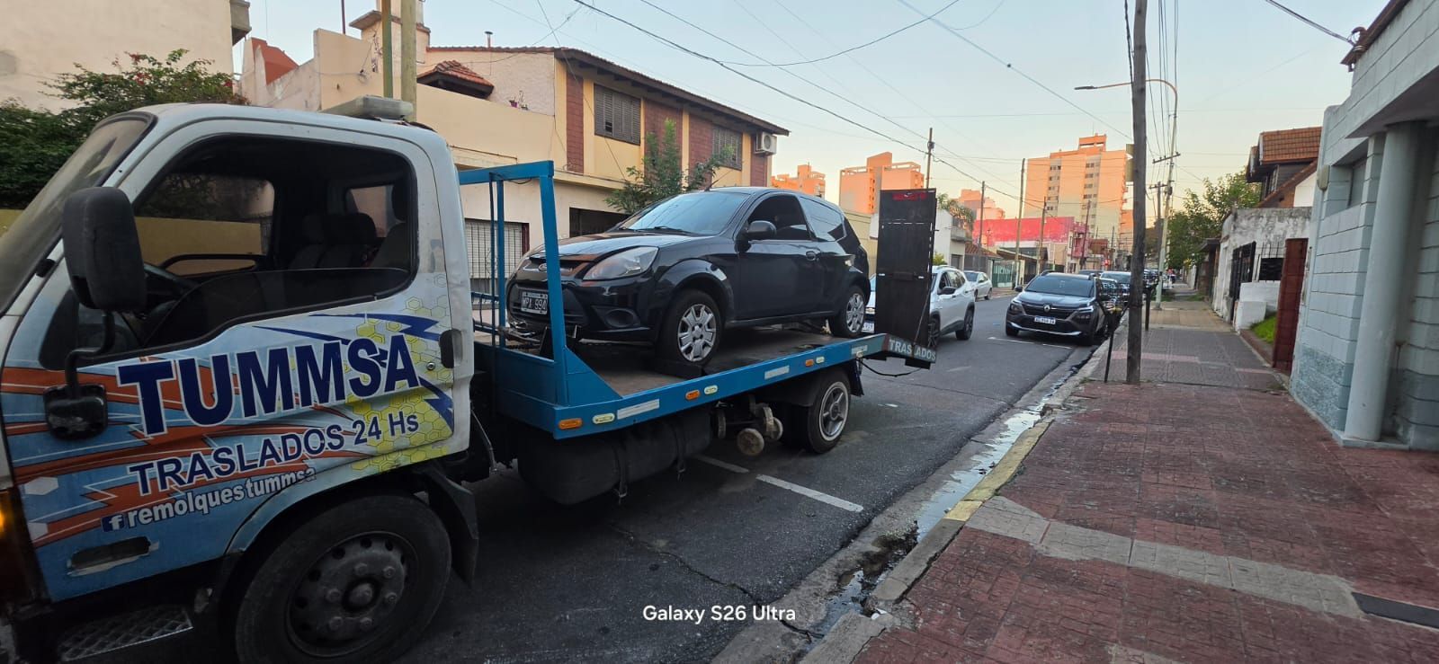 Una grúa transporta un coche azul en una calle de la ciudad, junto a vehículos y casas aparcados.