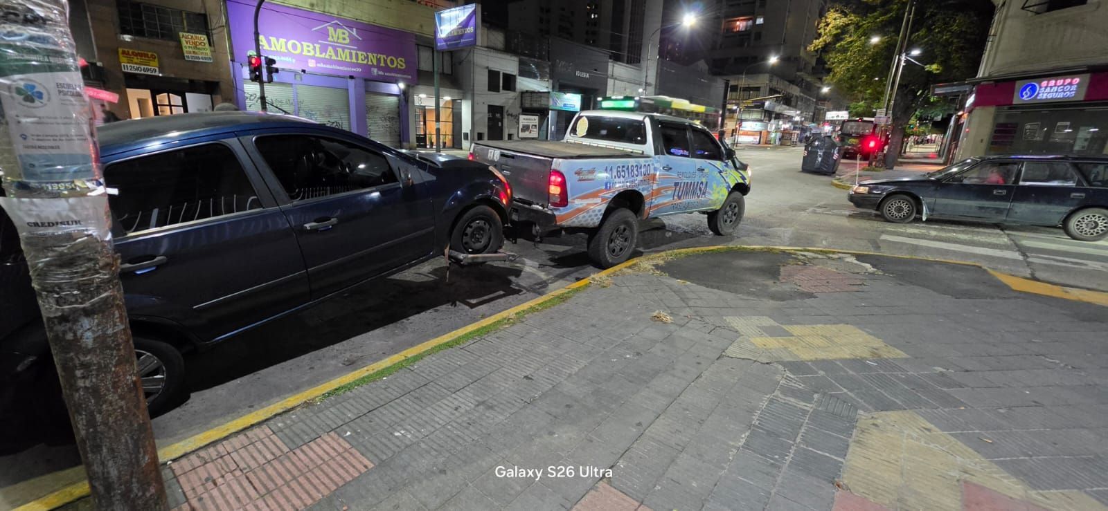 Escena nocturna en una calle con coches aparcados junto a un escaparate iluminado y marcas en la acera.