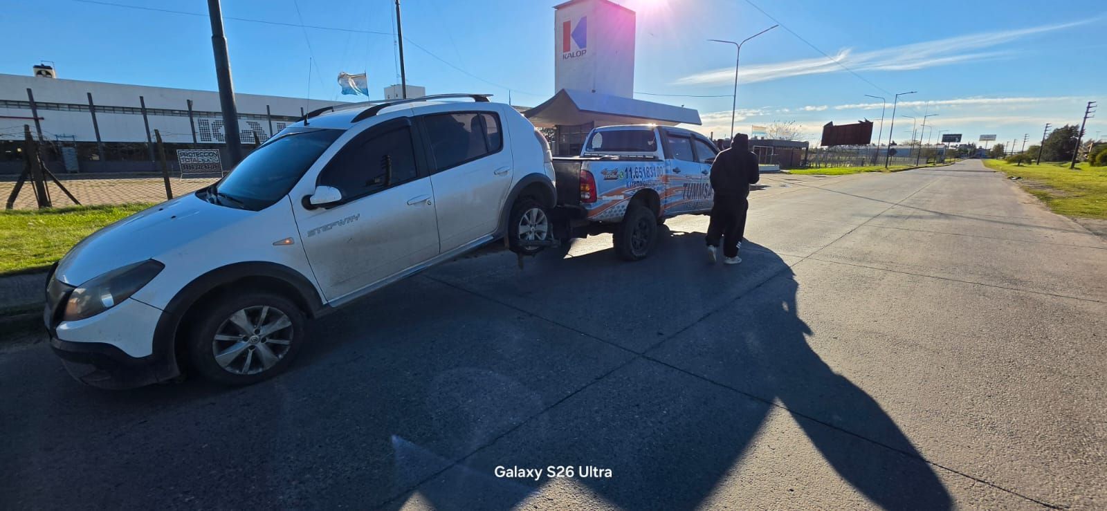 SUV plateado con enganche de remolque en una carretera soleada, con una persona de pie junto a él cerca de un remolque.