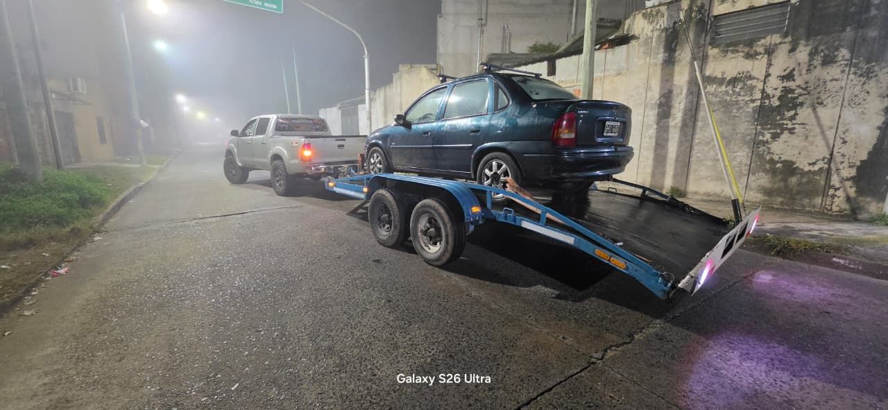 Un coche azul sobre un remolque por la noche, con otro vehículo detrás en una calle con niebla.