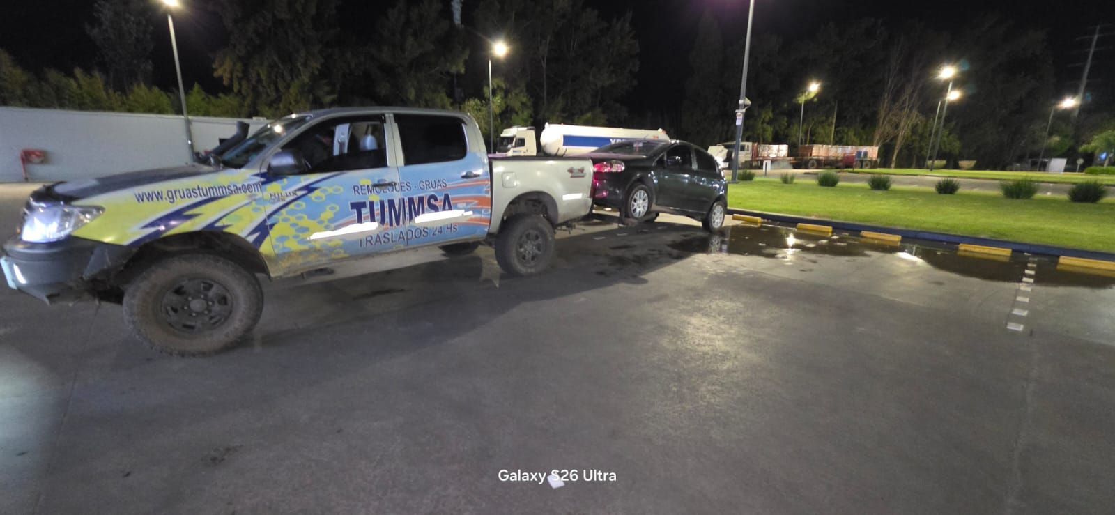 Camioneta policial y coche negro en un aparcamiento mojado por la noche con farolas y un charco en la acera.