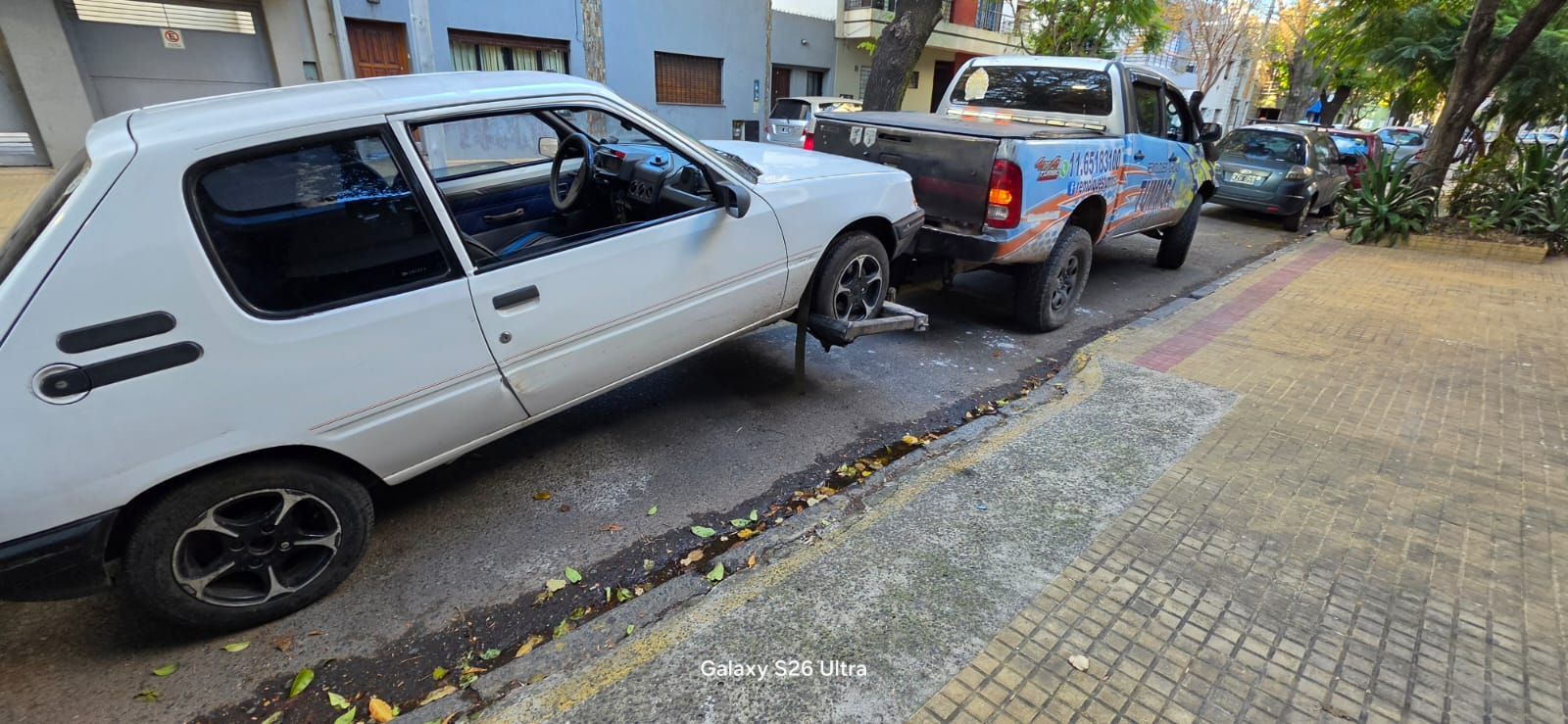 Camioneta blanca estacionada cerca de un auto negro en una calle estrecha junto a la acera.