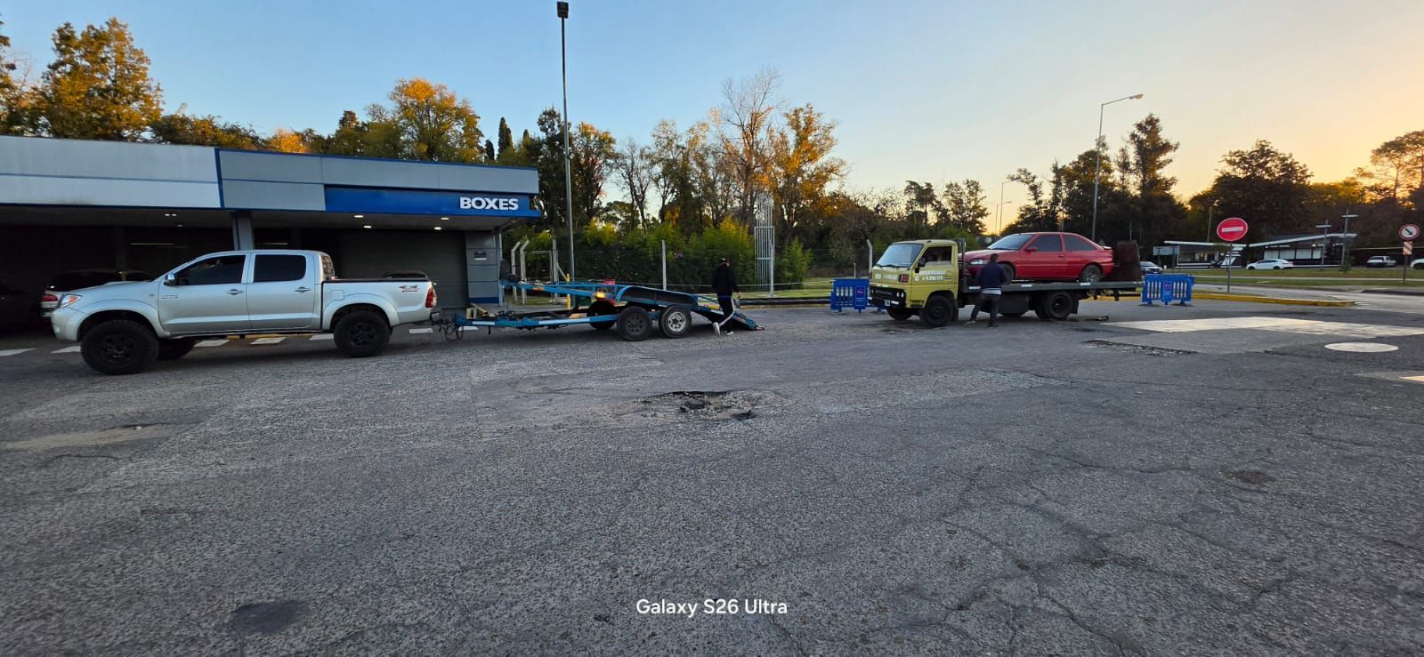 Estacionamiento con camioneta blanca, remolques y contenedores azules al anochecer.