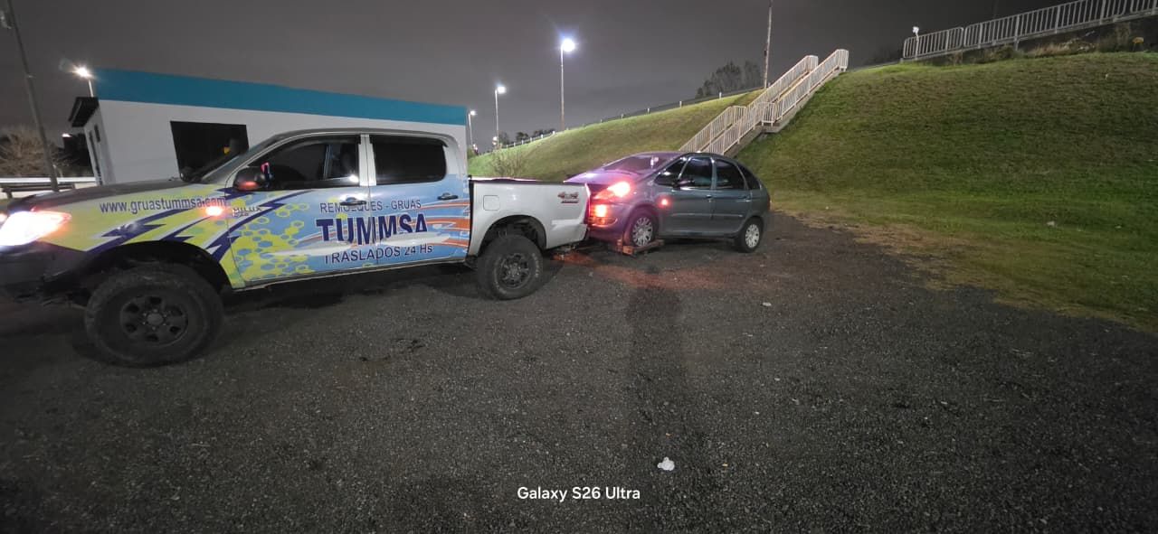 Un camión todoterreno y un coche se detienen en un camino de grava al anochecer, con colinas y farolas al fondo.