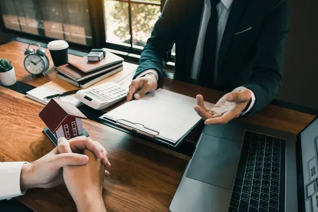 A man in a suit and tie is sitting at a desk talking to a woman holding a model house.