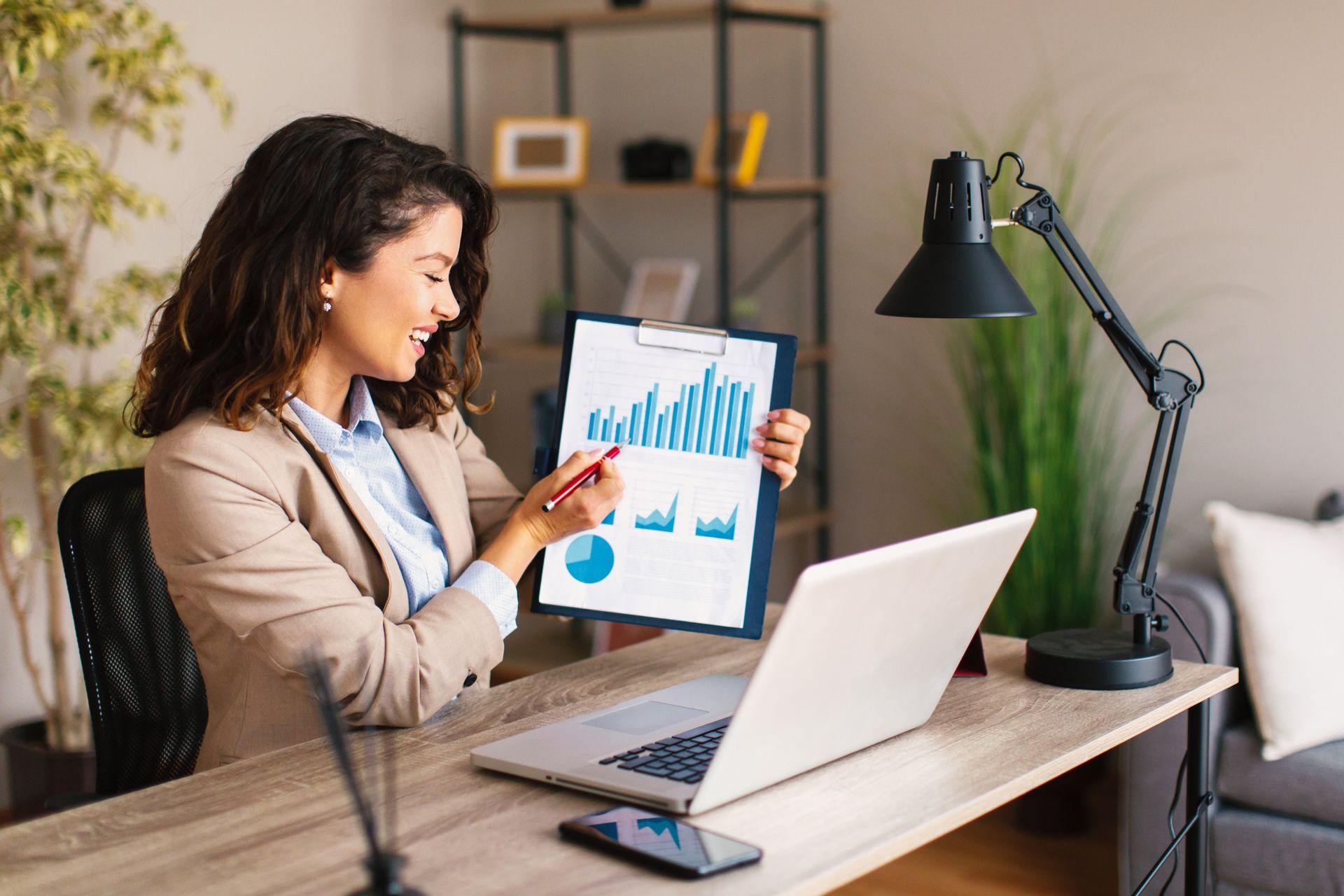 A woman is sitting at a desk with a laptop and holding a clipboard with a graph on it.