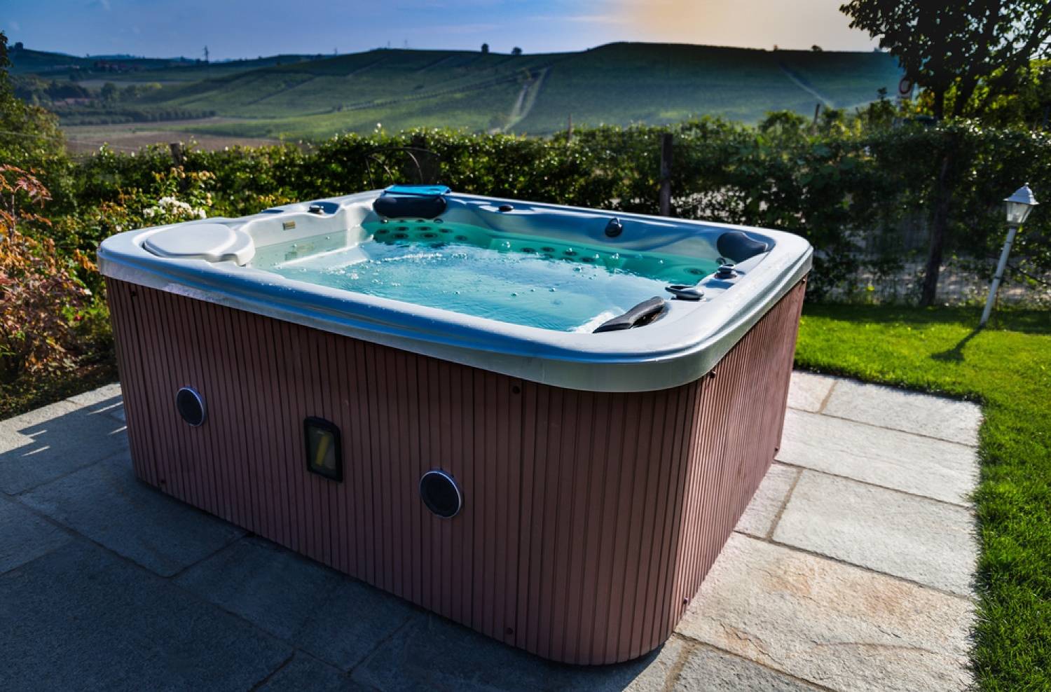 Outdoor hot tub filled with water on a patio, overlooking a green hillside and vineyard.