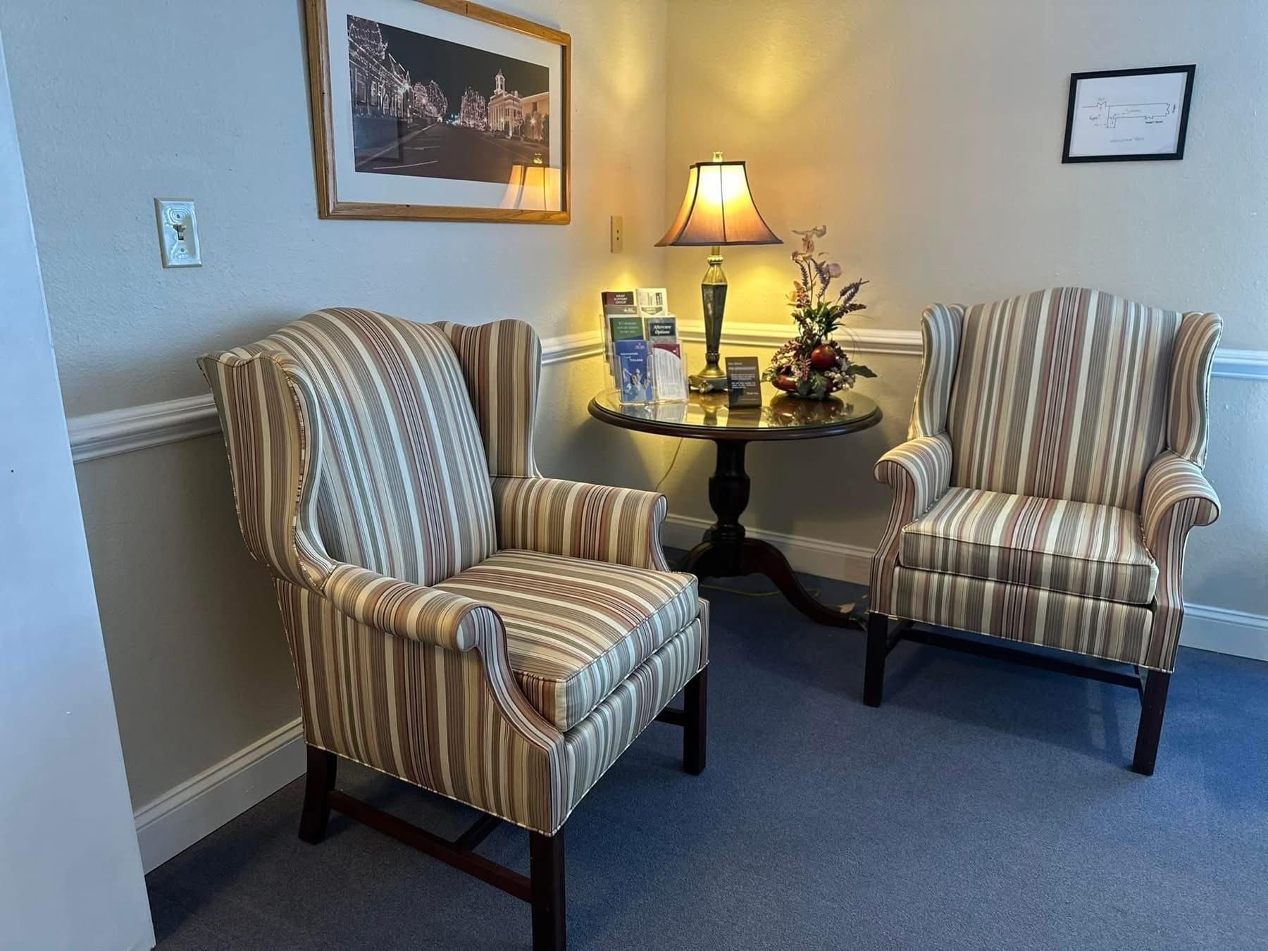 Two striped armchairs sit in a waiting area with a small table, lamp, and framed art.