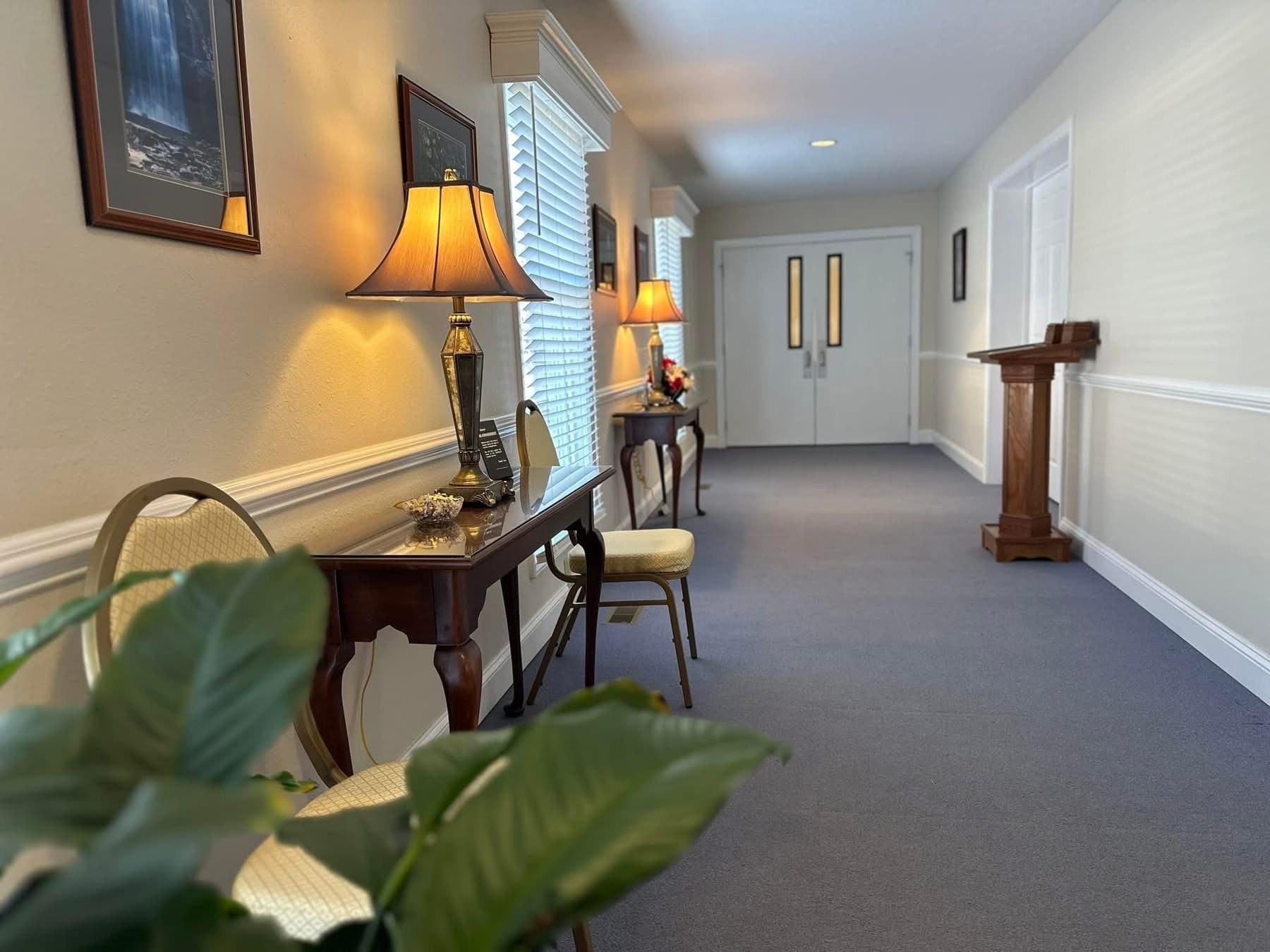Long hallway with blue carpet, adorned with lamps, tables, and a wooden podium. White walls and double doors at end.