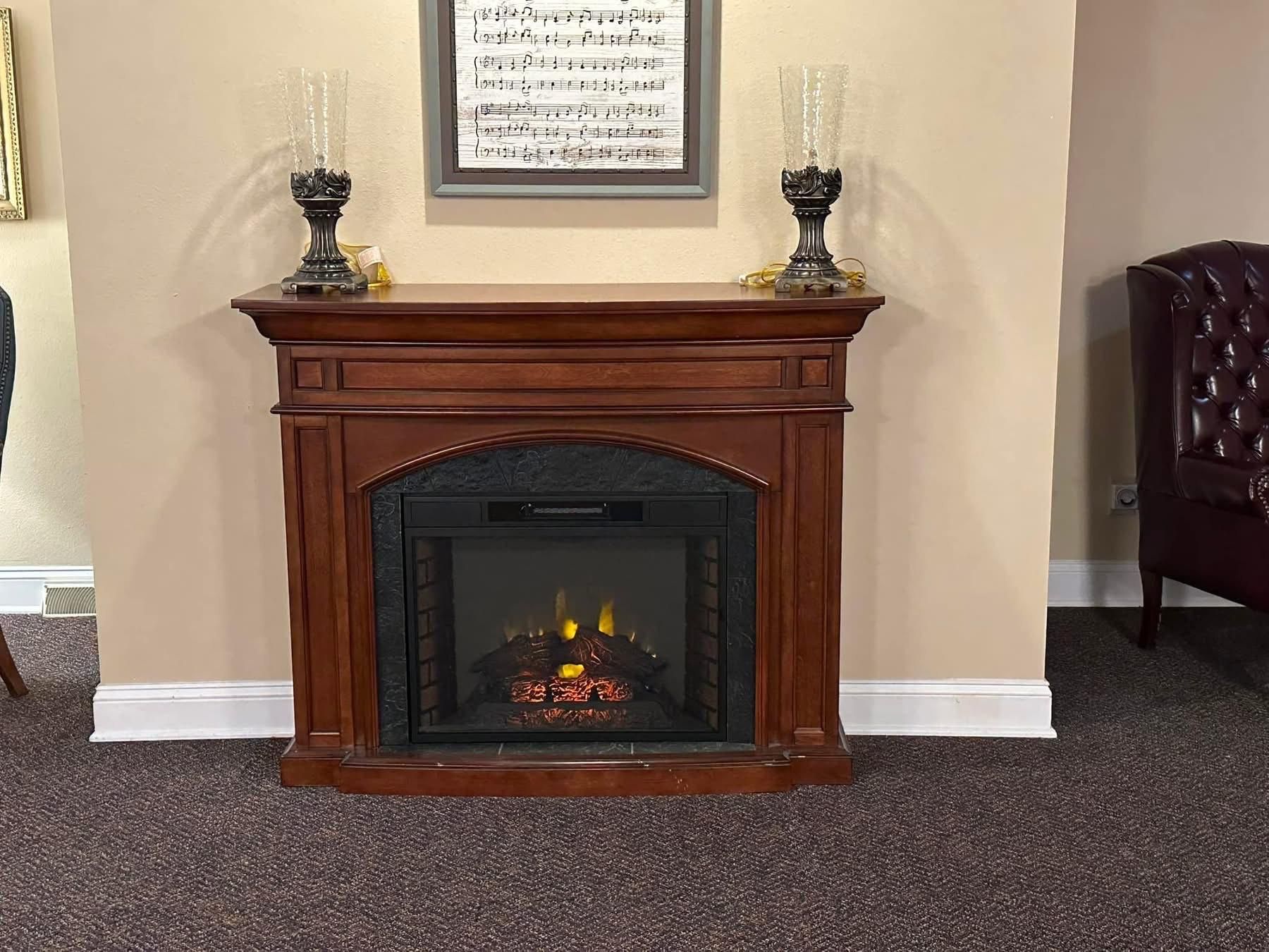 A wooden electric fireplace with flames, topped by candle holders and a framed musical score.