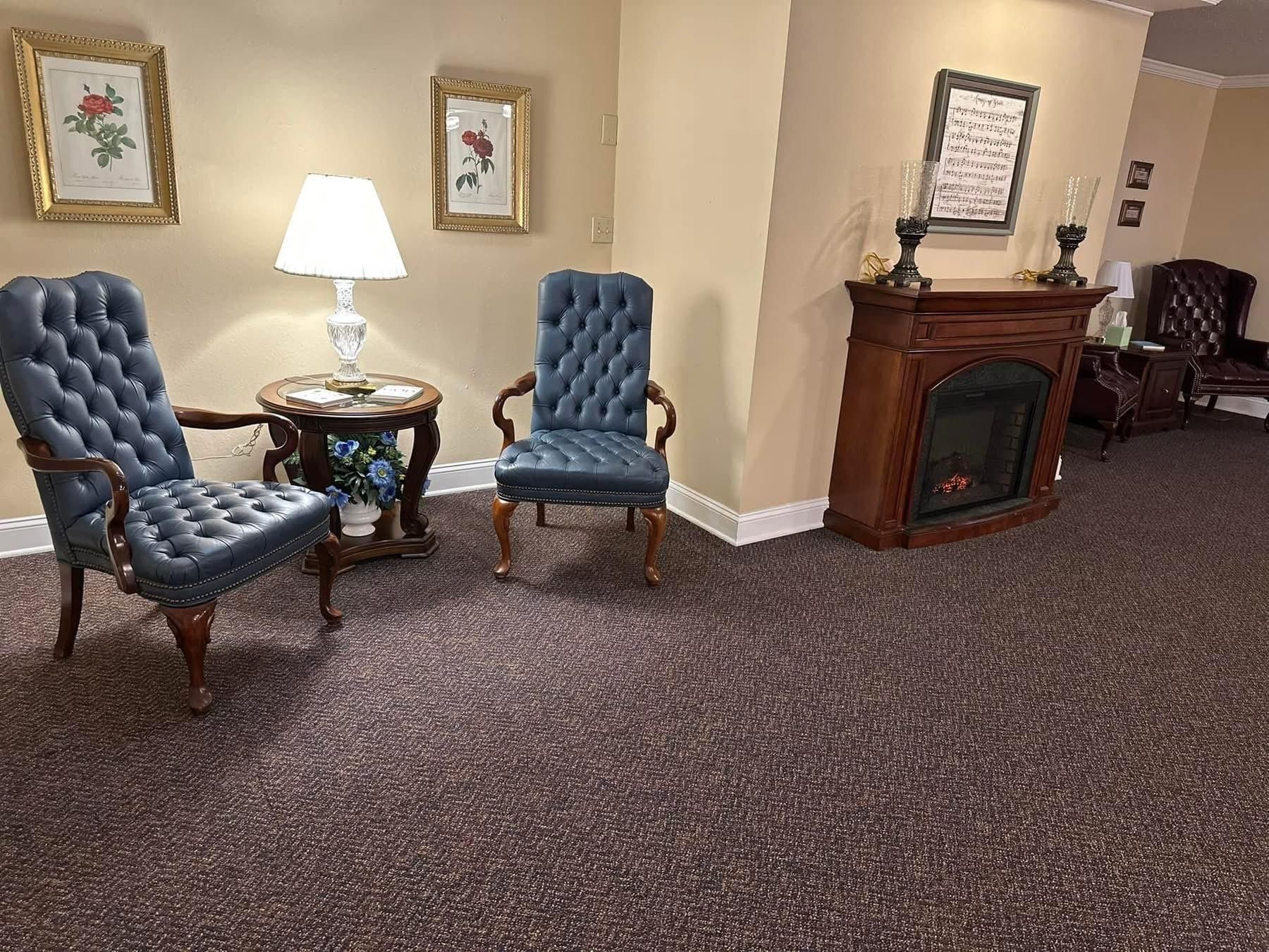 Two blue tufted chairs by a table with a lamp; fireplace and framed art in a room with brown carpet.