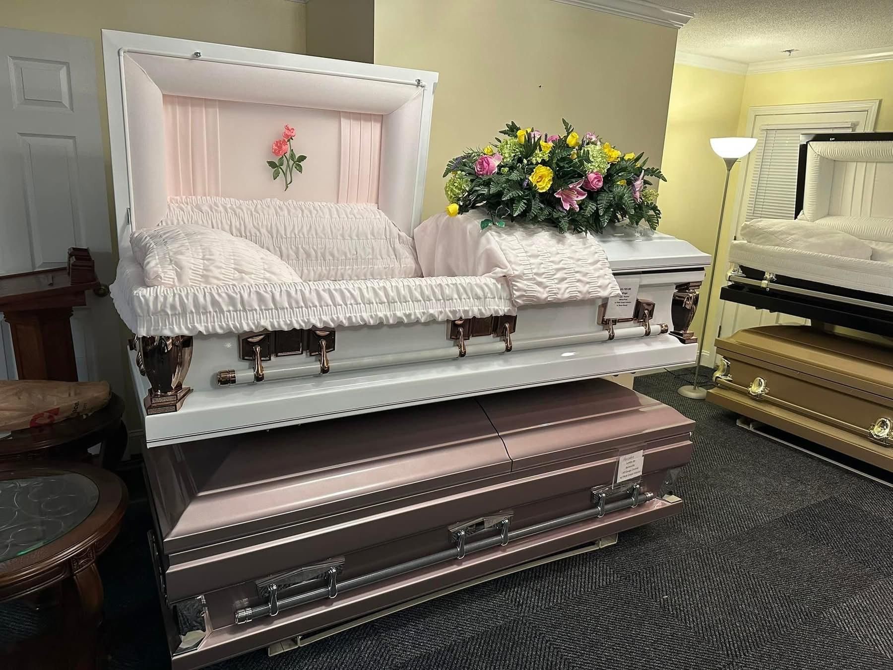 A display of caskets, including an open white one with pink interior and floral arrangement, in a store.
