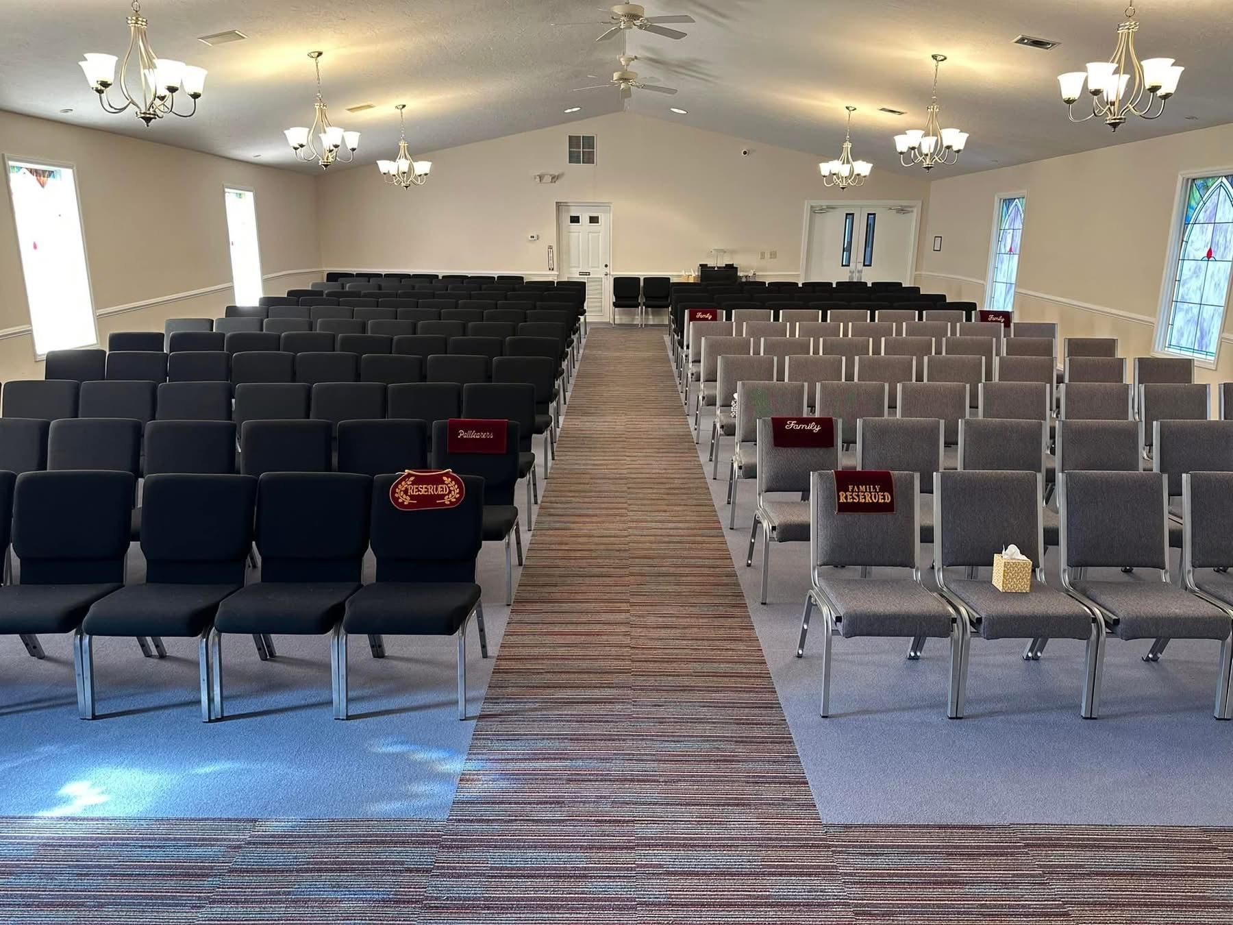 Interior of a large venue with rows of black and grey chairs, carpeted aisle, and a white door in the back.