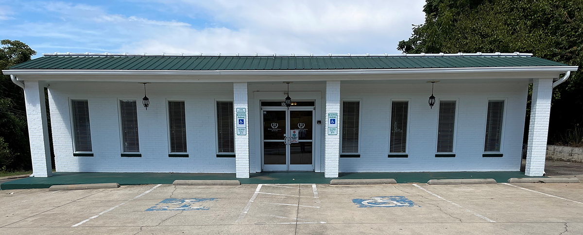 White building with green roof and trim, several windows and double doors, blue sky background.
