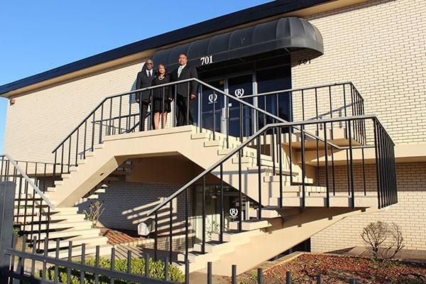 Three people stand outside a building with stairs. Black awning over the door.