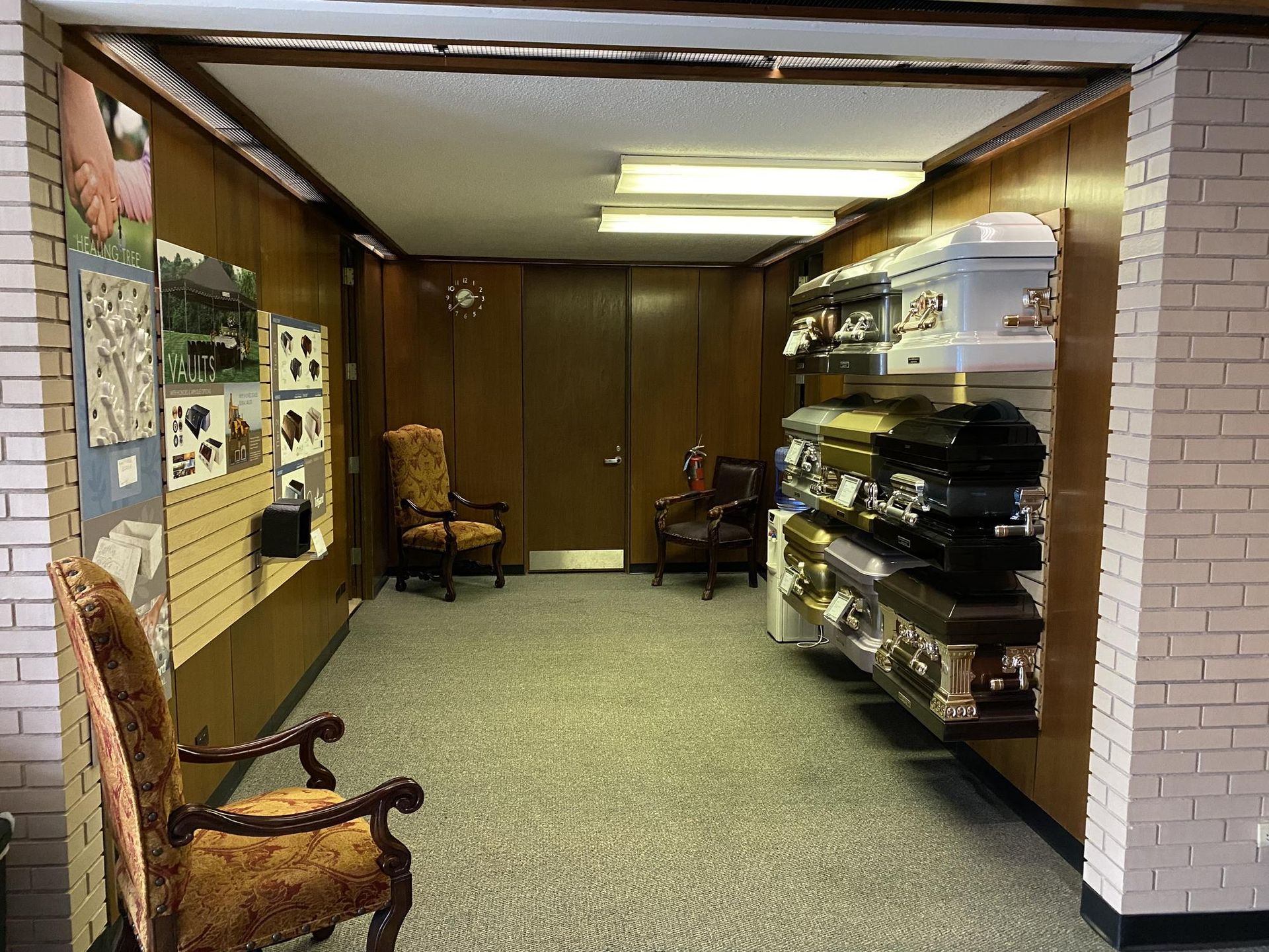 Interior of a funeral home displaying caskets. Two chairs and wall art are visible.