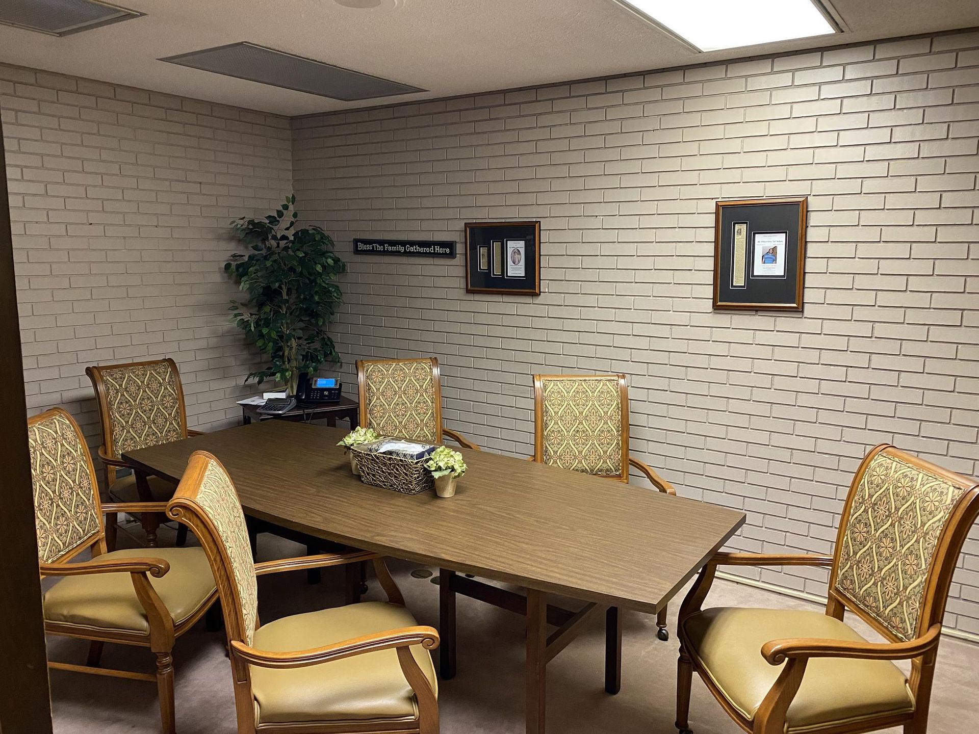 Meeting room with a brown rectangular table, patterned chairs, faux brick walls, and a plant.