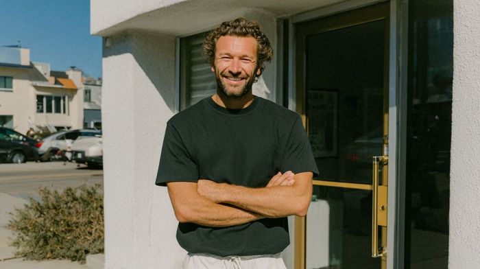 Man with crossed arms smiles in front of a building with a door, sunny day.