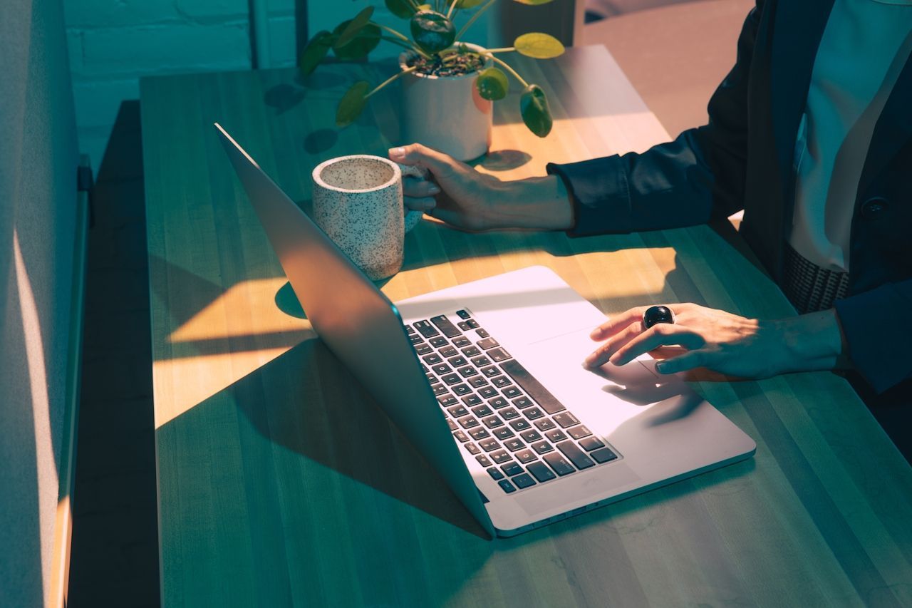 Person typing on laptop, holding mug, near potted plant on a wooden desk bathed in sunlight.