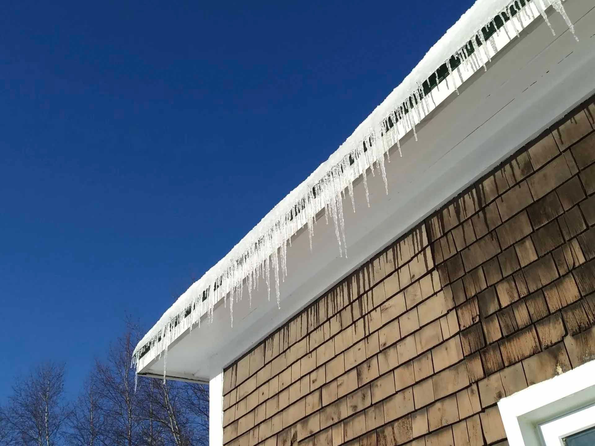 Des stalactites de glace pendent du toit blanc d'un bâtiment aux bardeaux bruns, sur fond de ciel bleu.