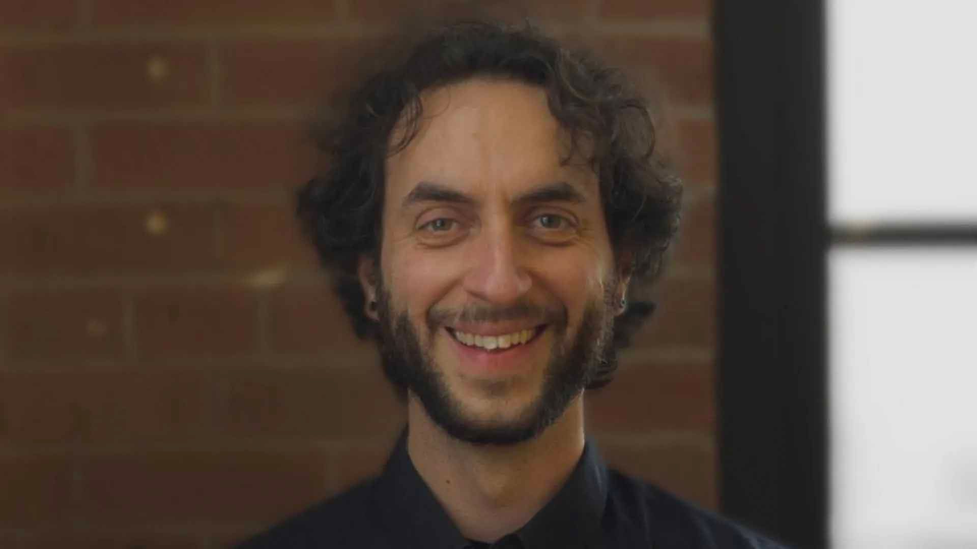 Un homme aux cheveux bouclés et à la barbe sourit devant un mur de briques.