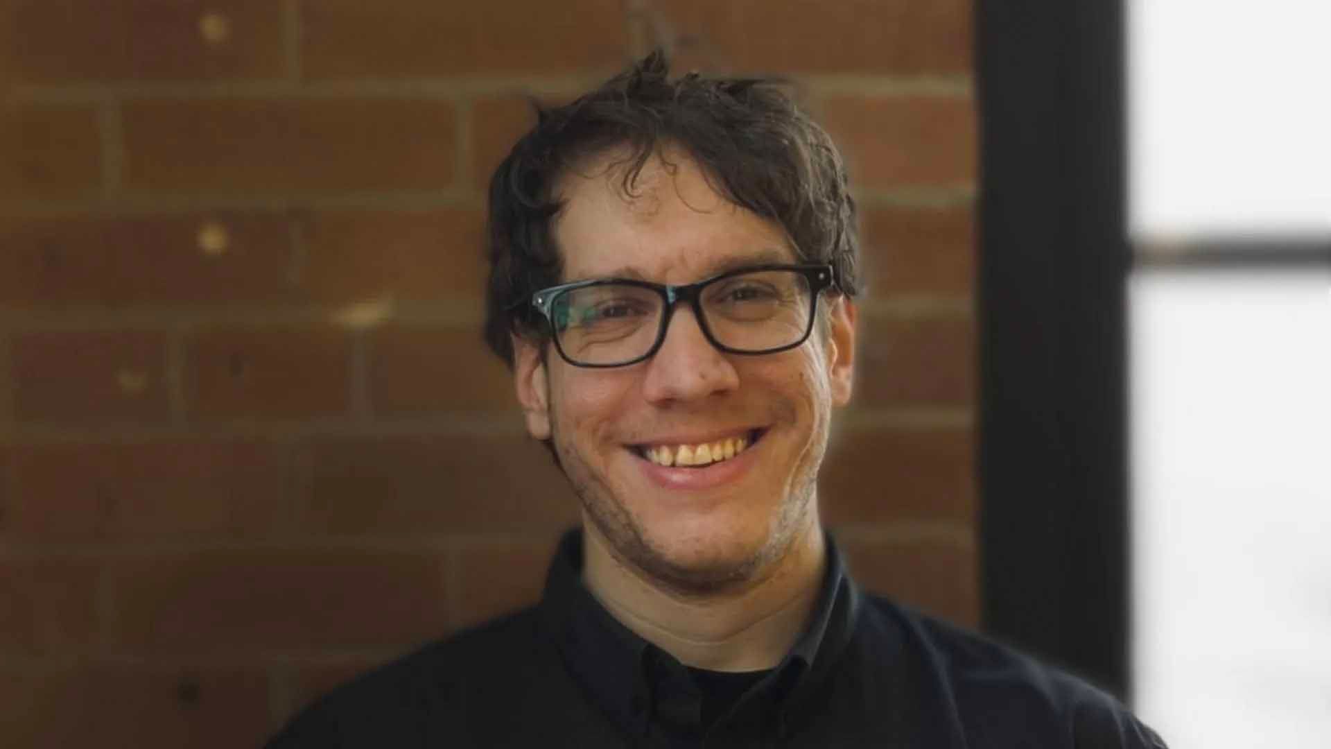 Un homme à lunettes sourit devant un mur de briques.