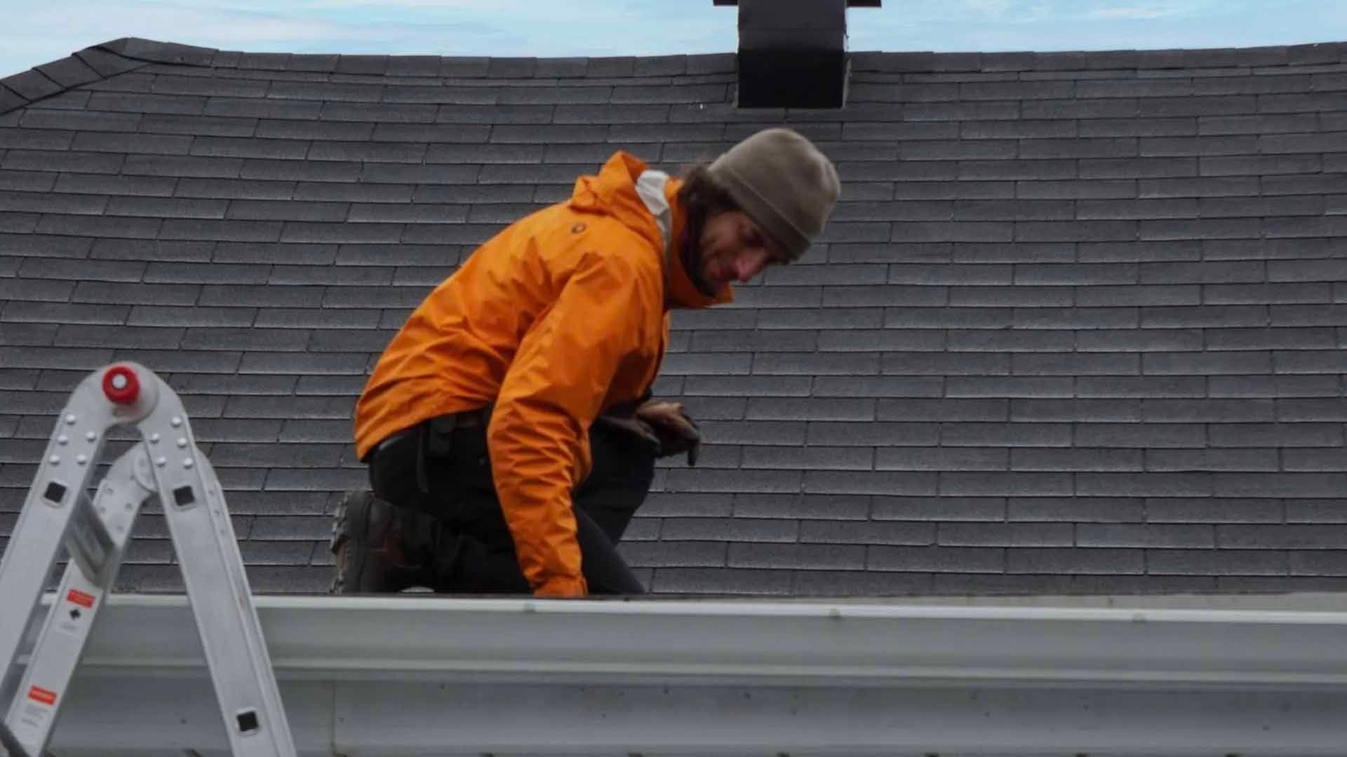 Une personne en veste orange inspecte une gouttière sur un toit. Toiture grise et ciel nuageux.