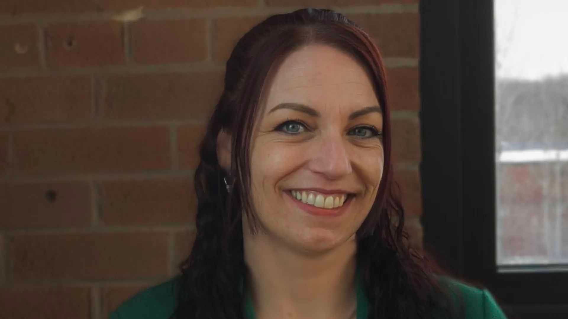 Une femme aux cheveux auburn sourit, regardant l'objectif, devant un mur de briques et une fenêtre.