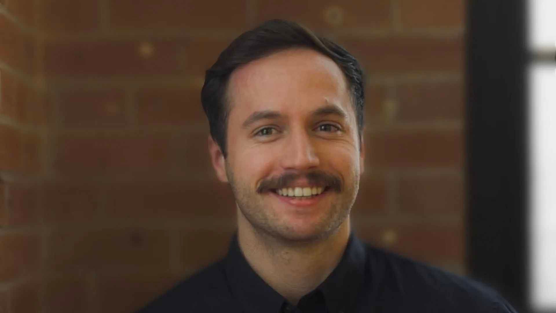 Un homme à moustache sourit, appuyé contre un mur de briques.