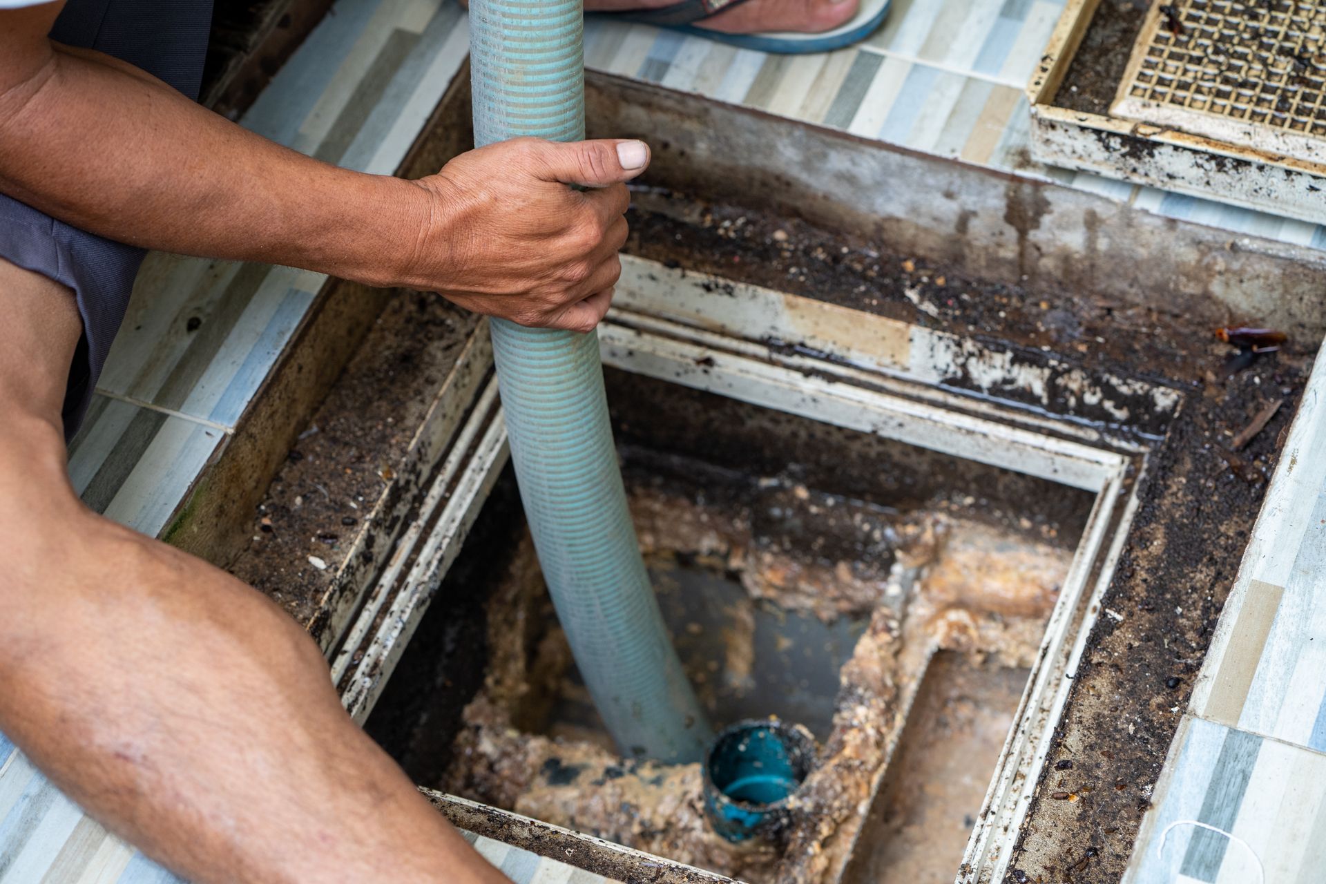 Man using a hose to clean a dirty drain in a tile floor.
